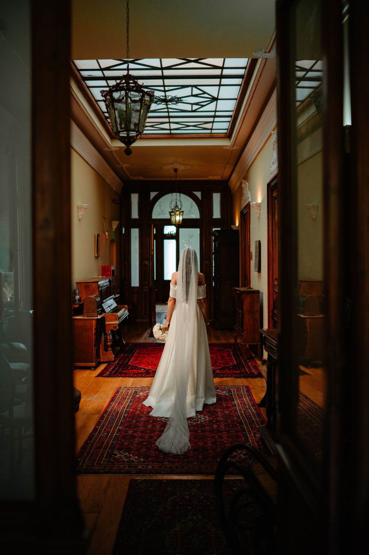 Bride in white wedding gown with veil standing in ornate hallway with stained glass ceiling.