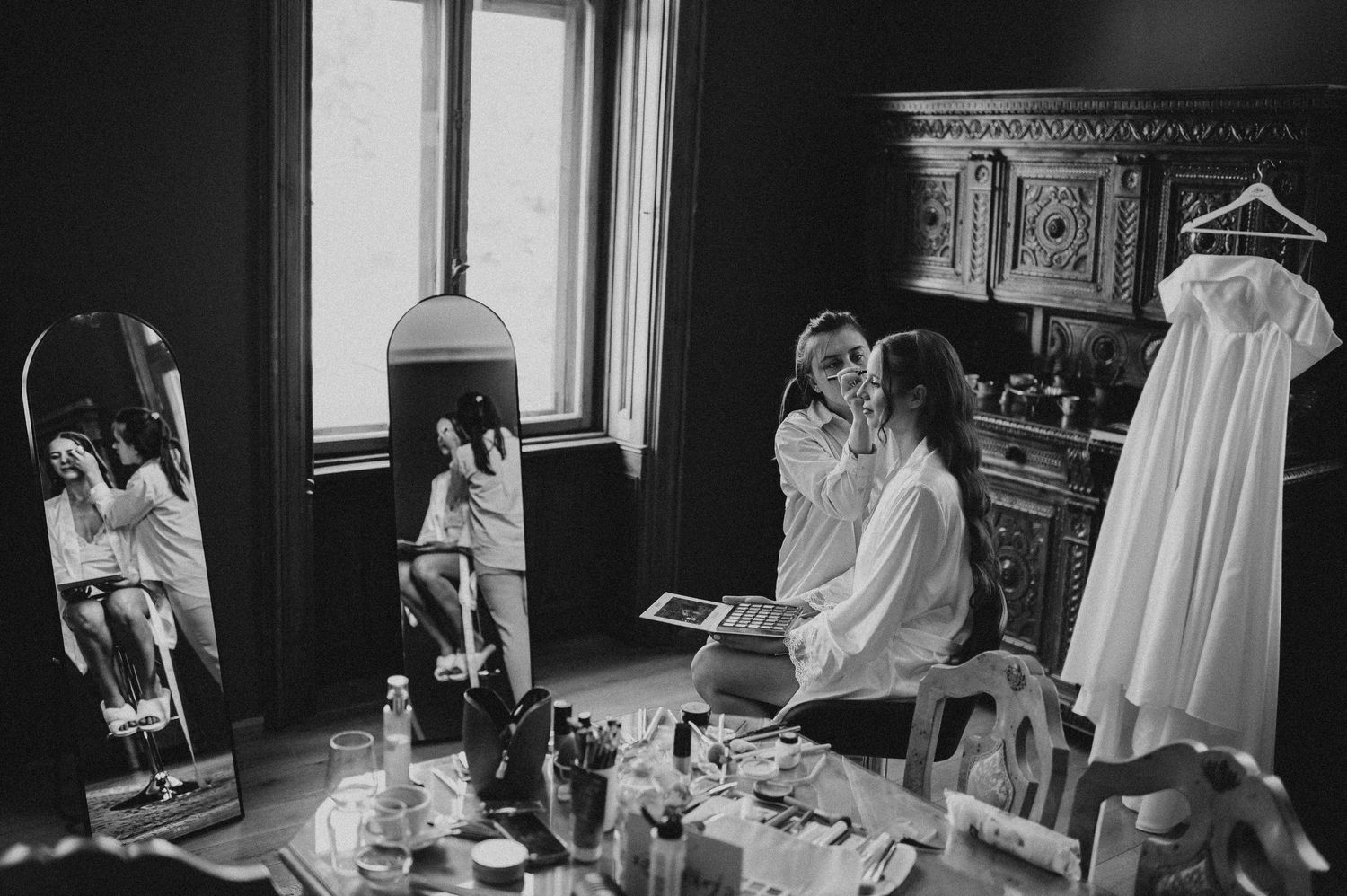 Black and white photo of bride getting makeup done in a ornate room with mirror reflection.