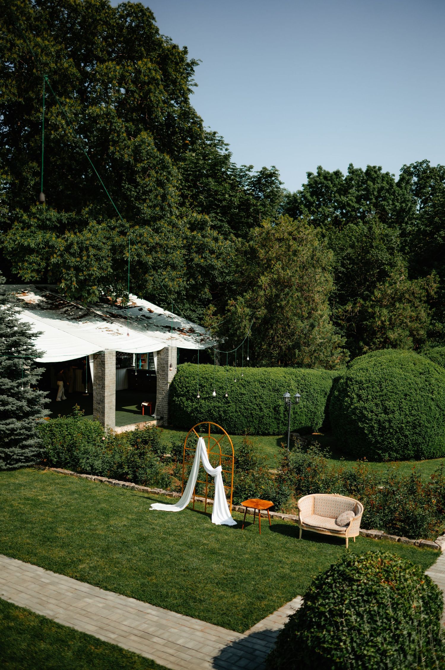 Outdoor wedding ceremony setup with golden arch, white draping, and manicured hedges in sunlit garden.