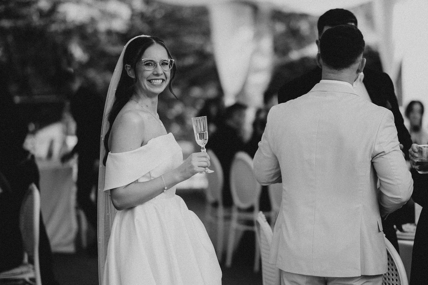 Smiling bride in off-shoulder gown holding champagne glass at outdoor wedding reception.