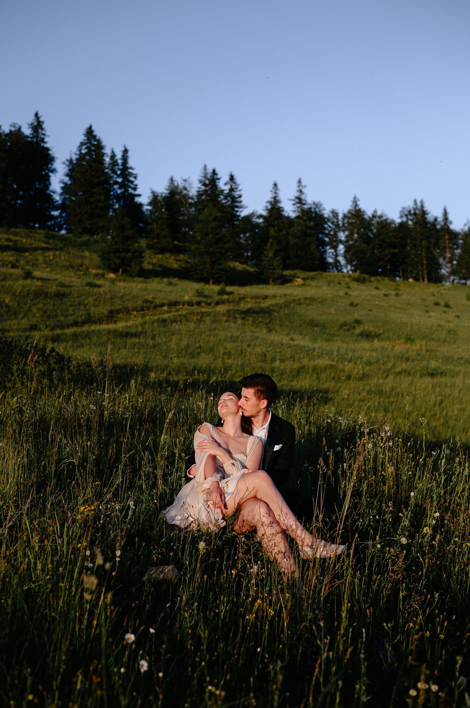 A bride and groom share a romantic kiss while sitting in a lush green meadow at golden hour.