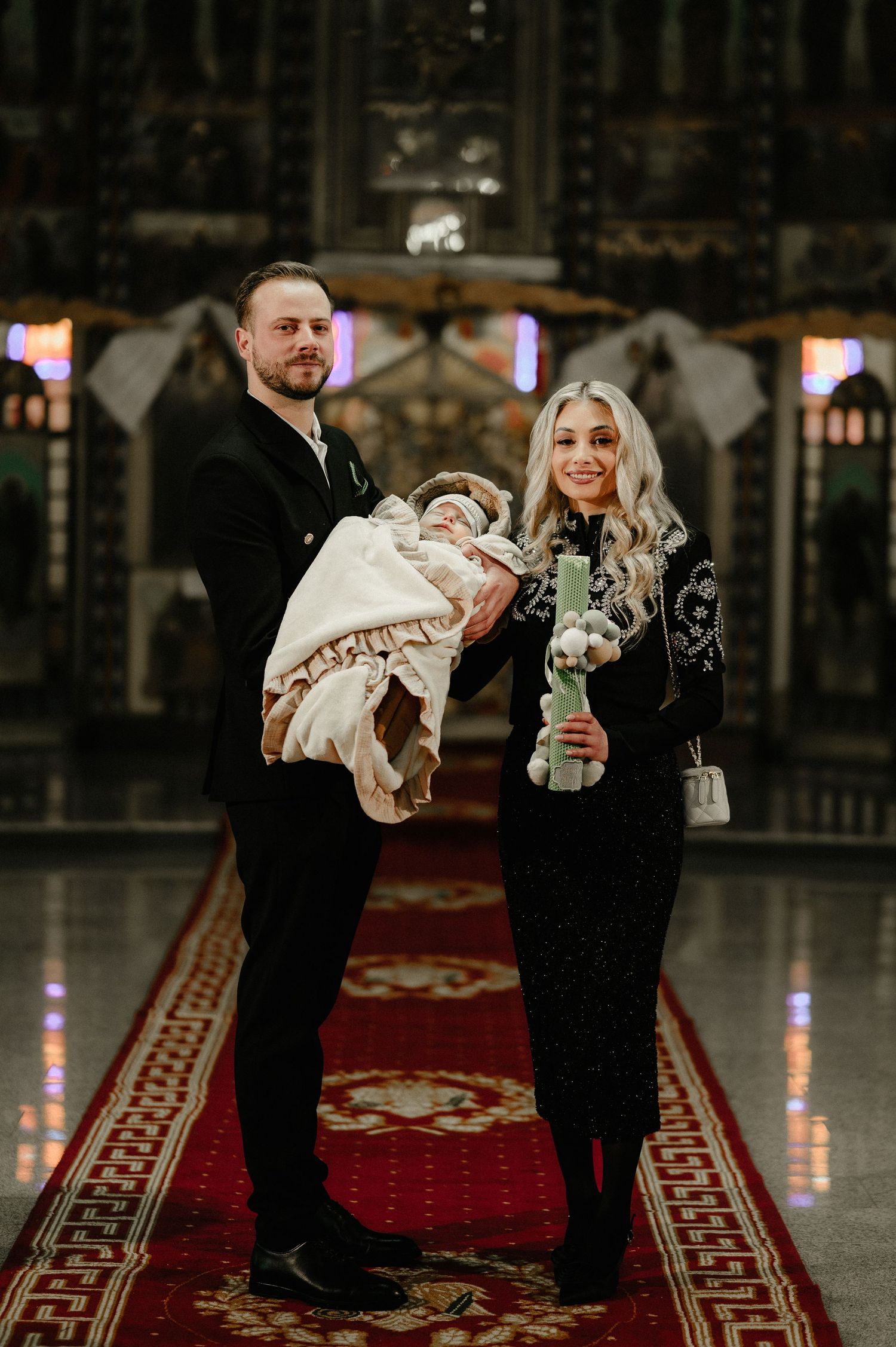 Couple in black formal attire holding newborn baby and baptism candle inside ornate church.
