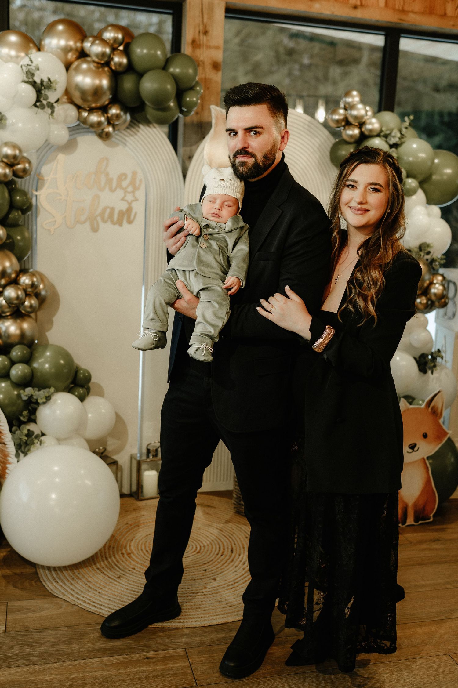 A couple in black outfits holding their newborn baby at a woodland-themed christening party with green and gold balloons.