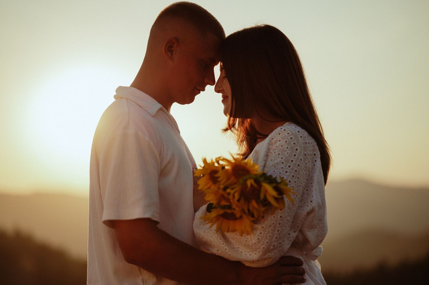 Romantic couple touching foreheads holding sunflowers in silhouette against bright sunset.