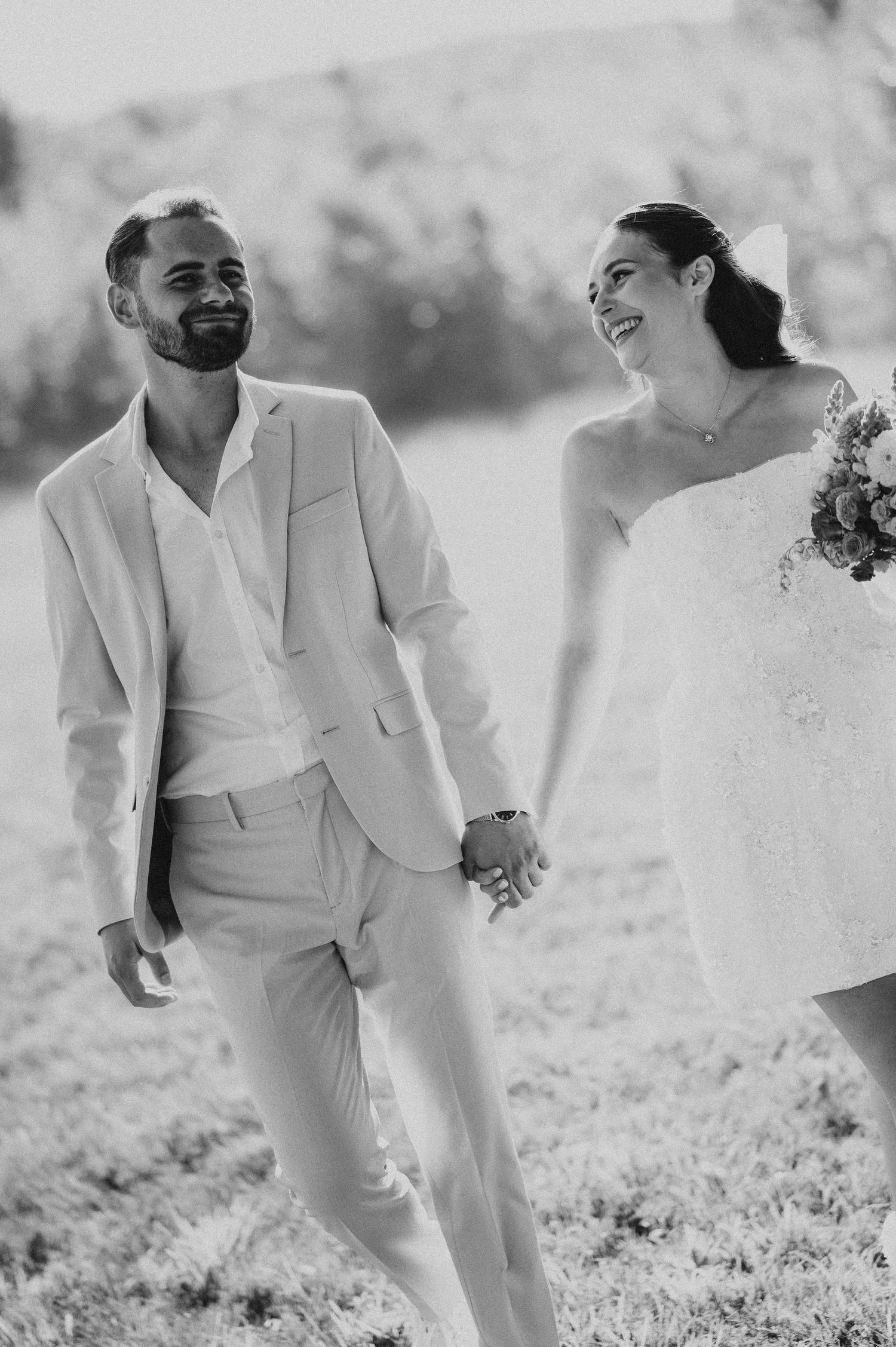 Black and white photo of smiling newlyweds holding hands and walking through a sunlit grassy field.