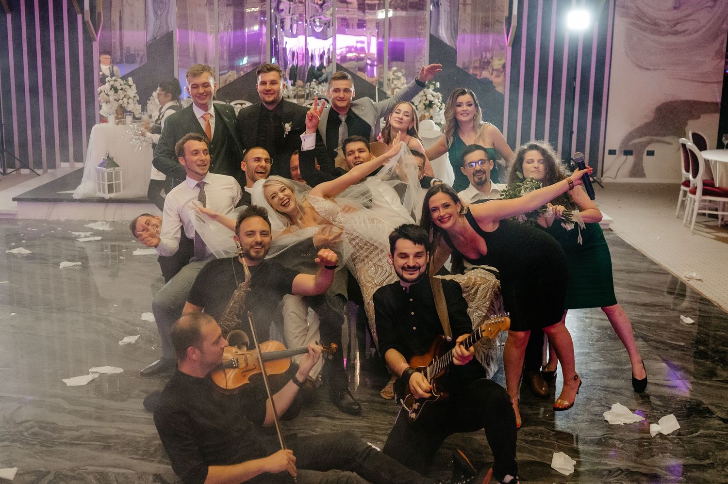 Wedding party group photo with musicians holding violins celebrating on the dance floor at a reception.