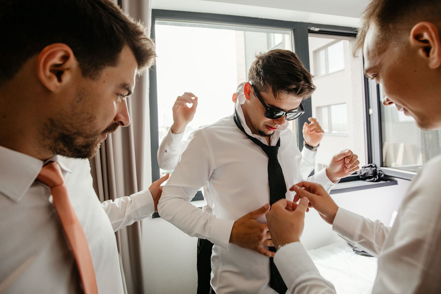 Groomsmen assist groom in black suit with cufflinks and tie during wedding morning preparations.
