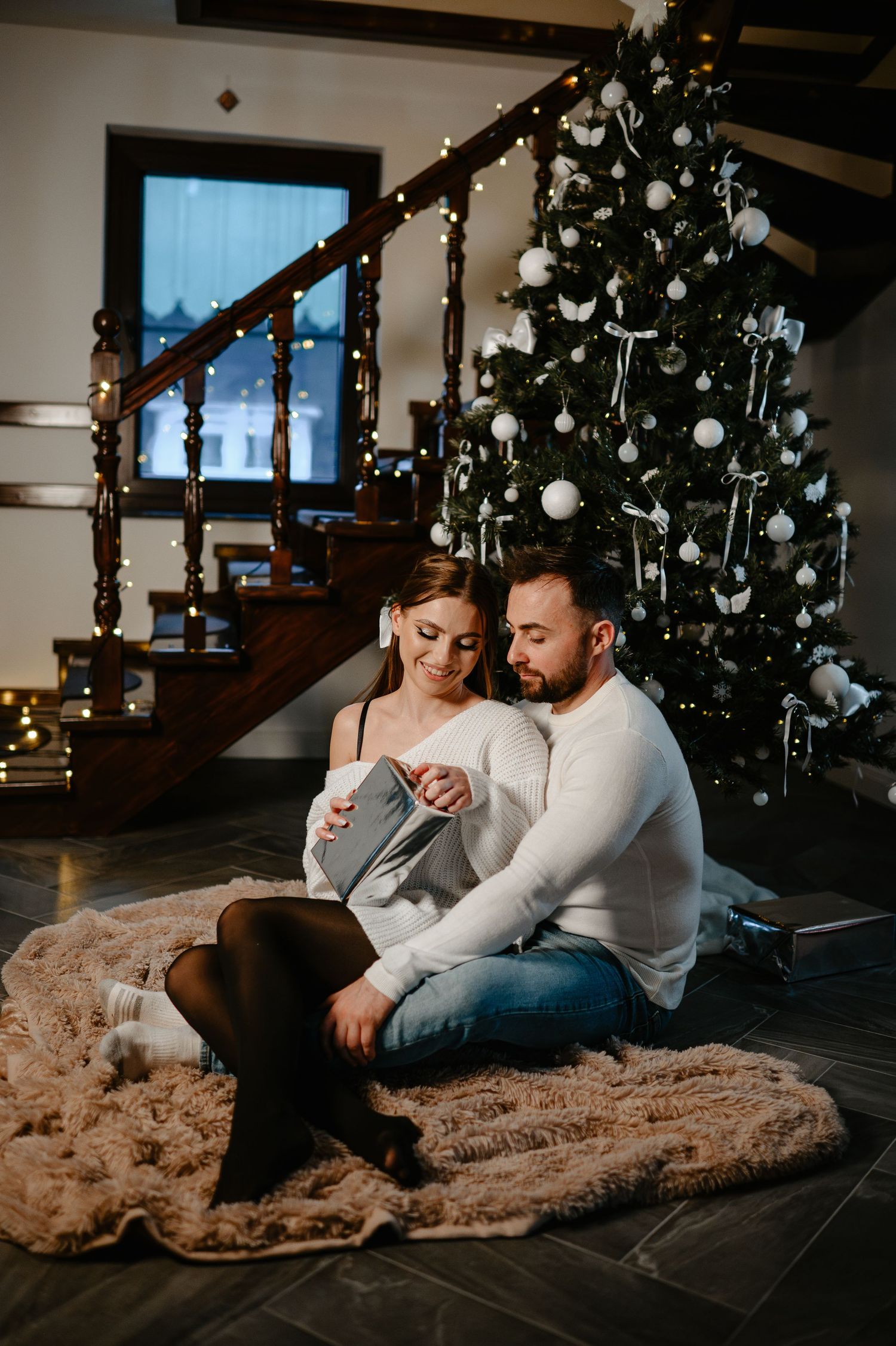 Couple sitting on a fur rug in front of a decorated Christmas tree in a cozy home.
