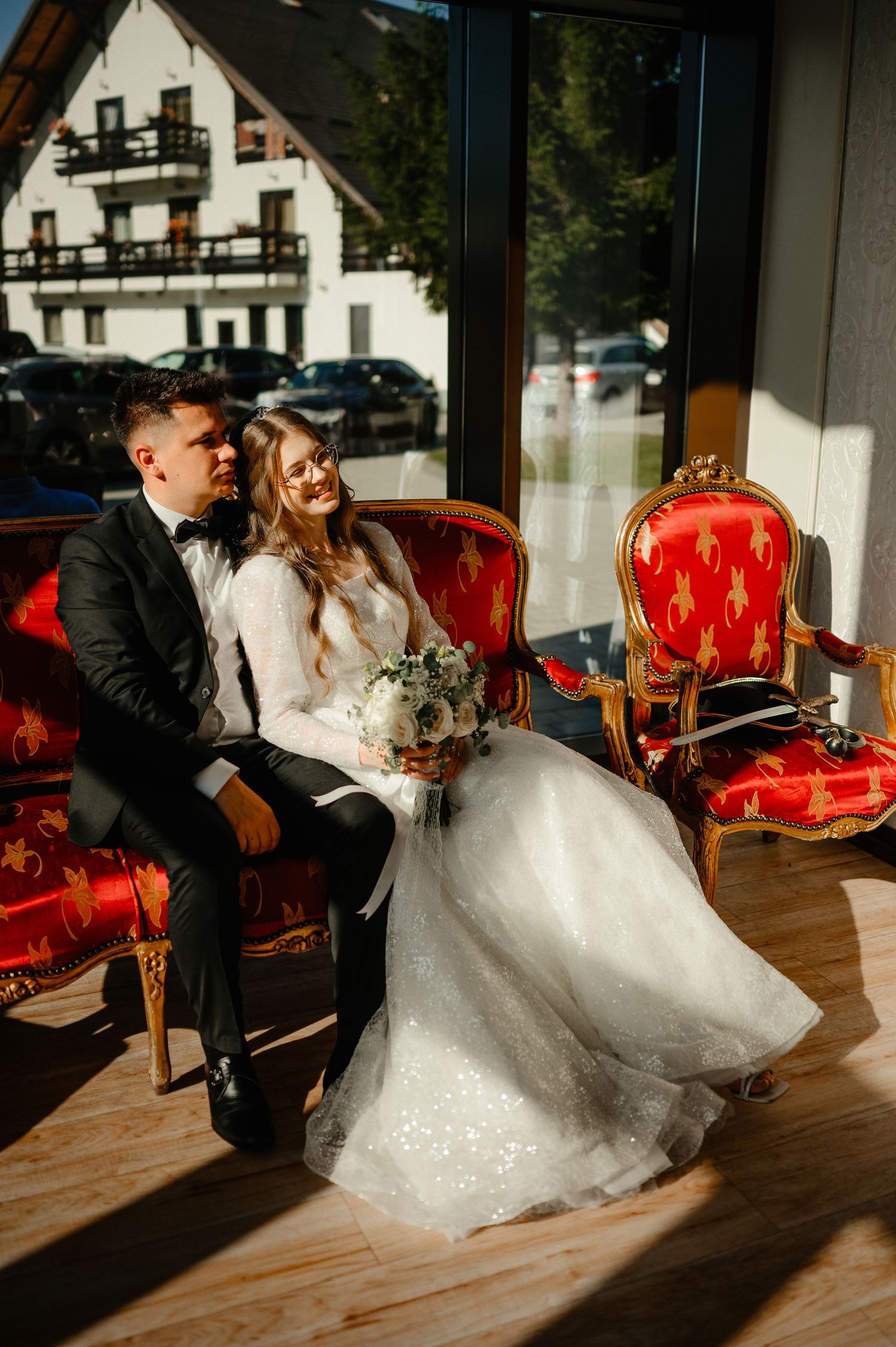 Bride and groom sitting together on a red vintage sofa by a large sunlit window.