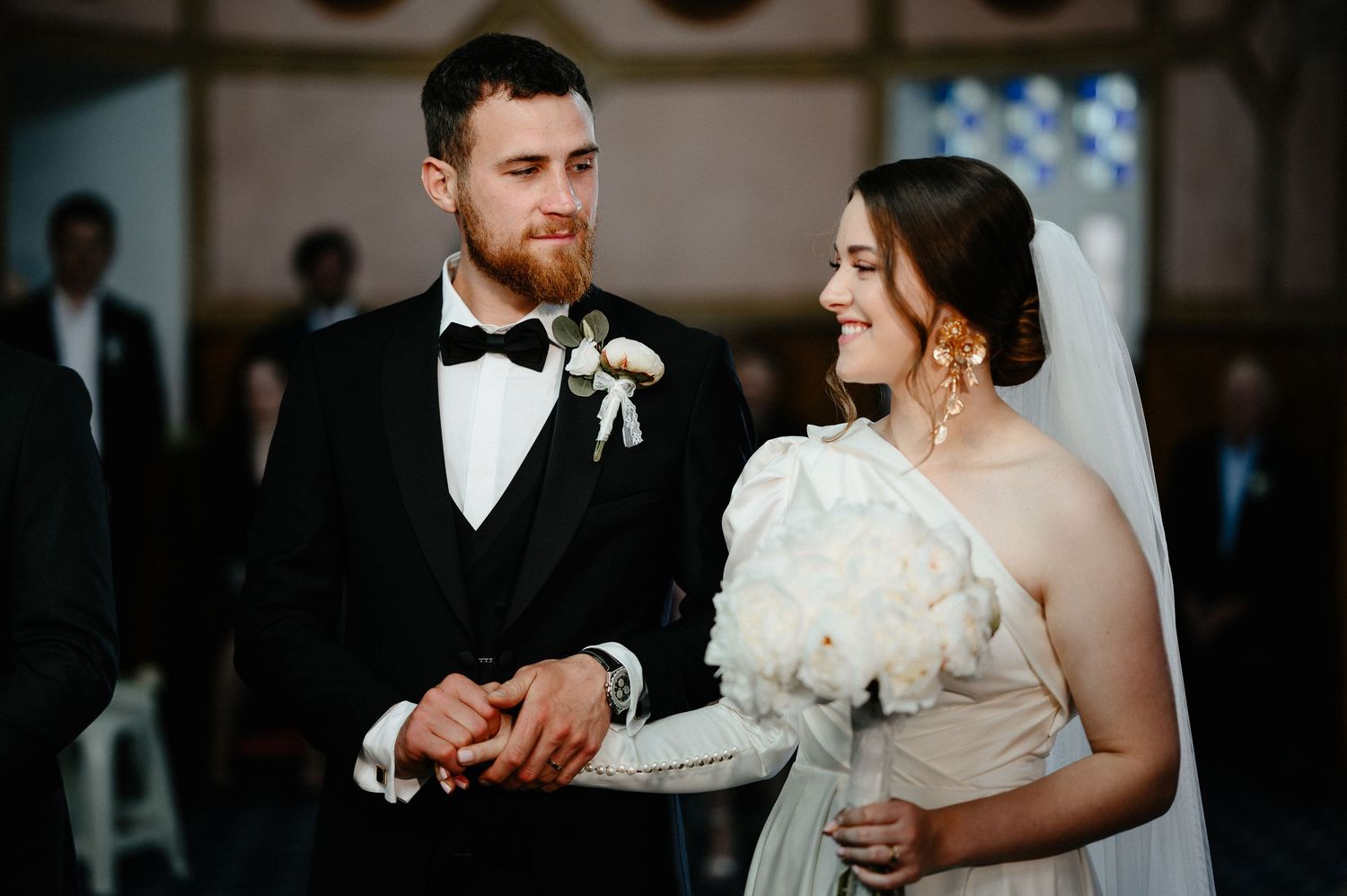 Bride and groom smiling at each other during wedding ceremony, bride holding white bouquet.
