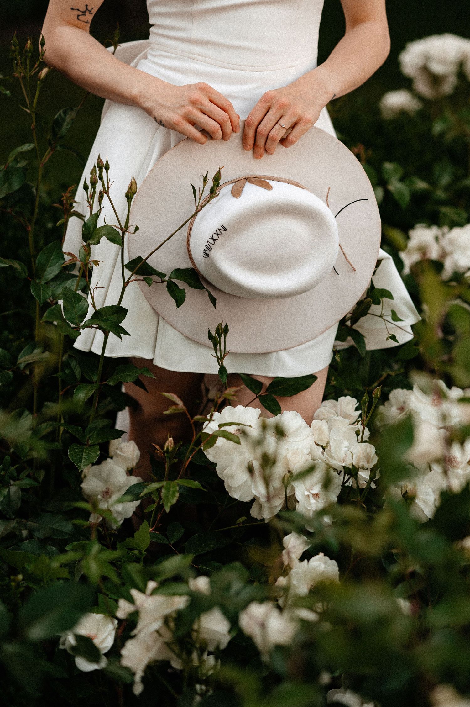 Woman in white dress holding a wide-brim hat surrounded by blooming white roses.