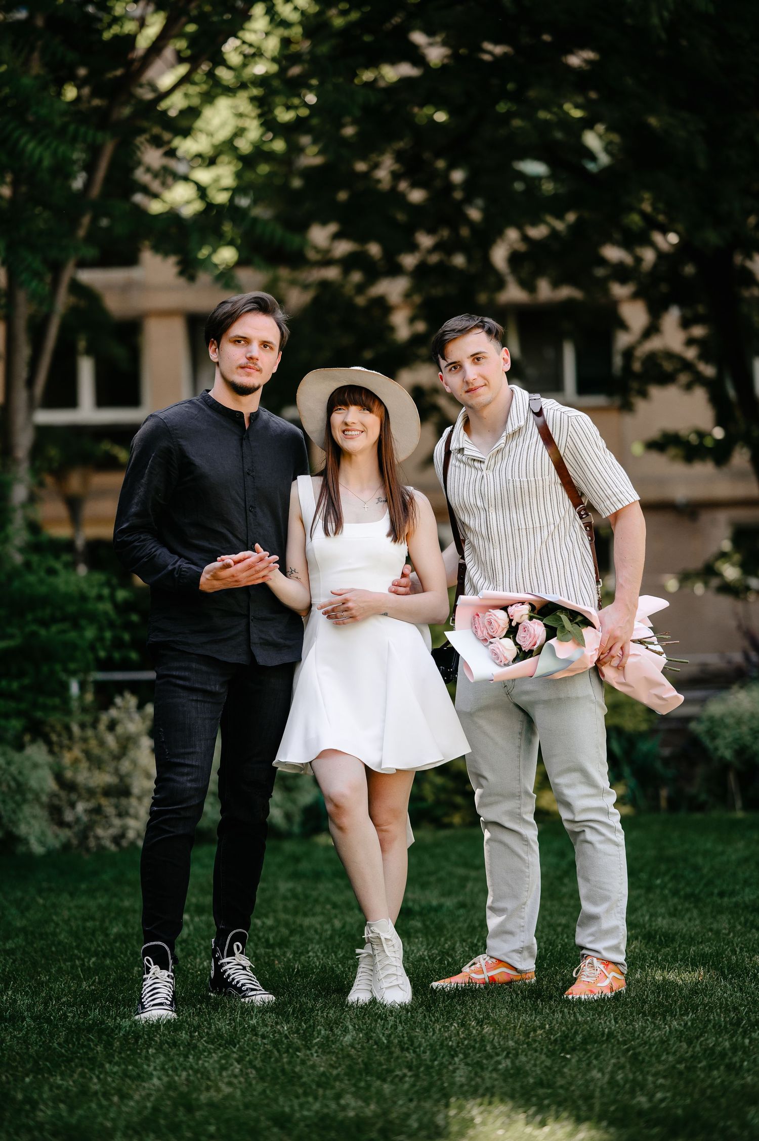 Three young friends posing in a green park, woman in white dress and hat flanked by two men, one holding pink flowers.