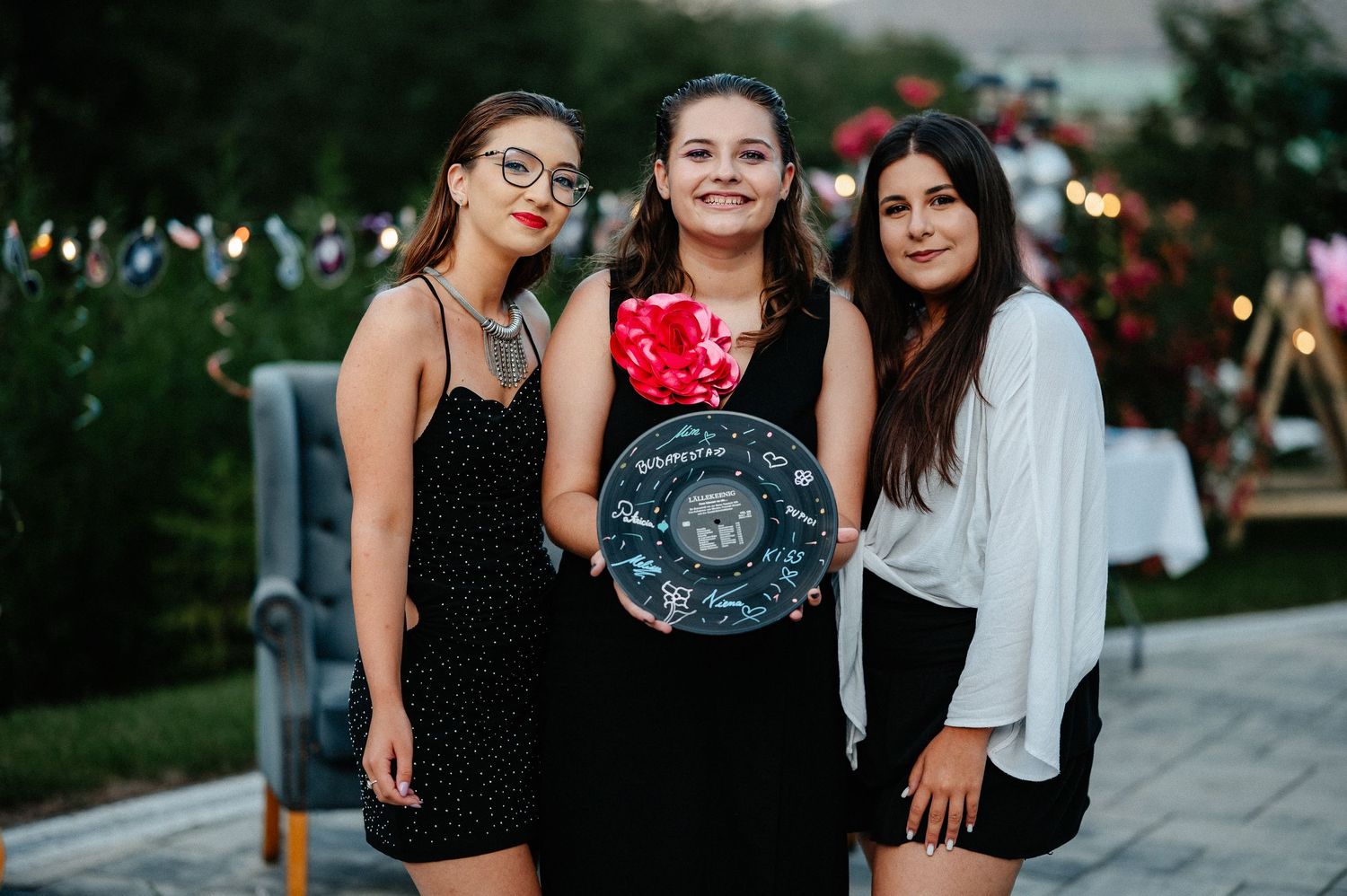Three young women posing together outdoors, holding a decorated vinyl record at a birthday party.
