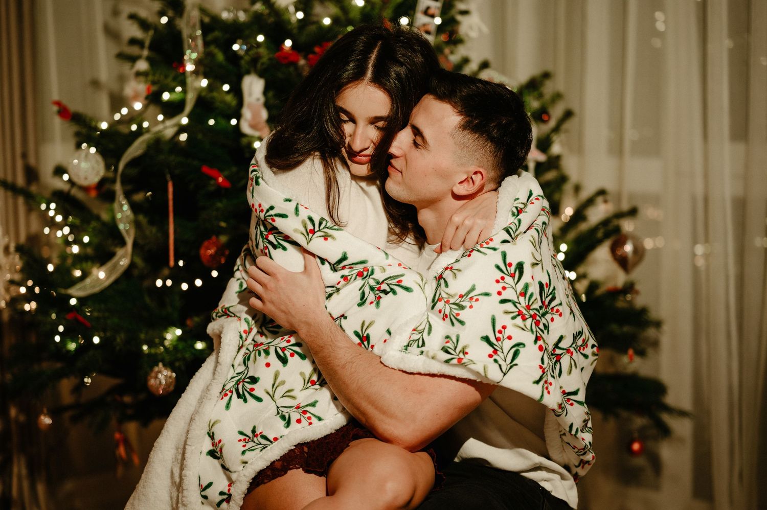 Couple cuddling under a floral blanket in front of a decorated Christmas tree.