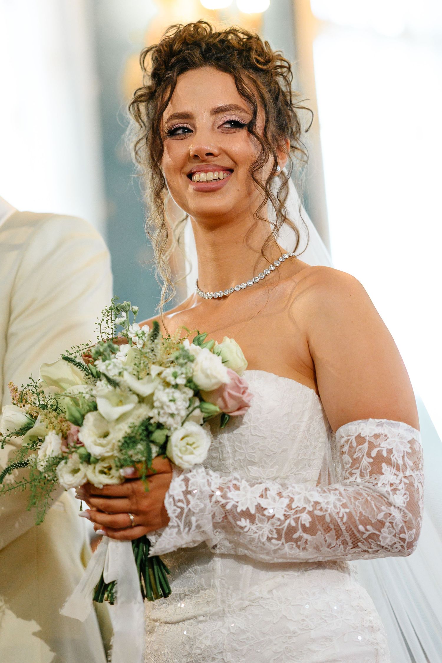 Smiling bride in white strapless lace gown holds bouquet of white and pink roses, wearing diamond necklace and veil.