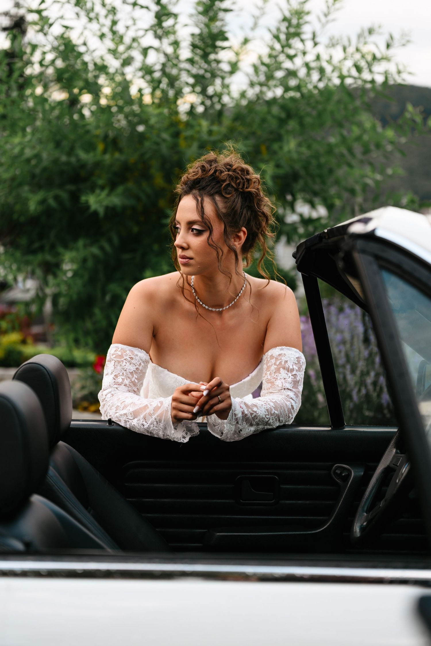 Elegant woman in white lace off-shoulder top leaning on a convertible car outdoors.