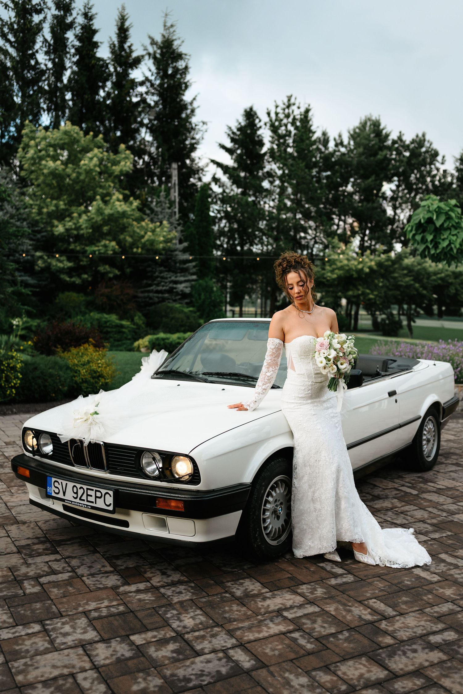Bride in white lace gown holding bouquet poses beside a classic white BMW convertible decorated for a wedding.
