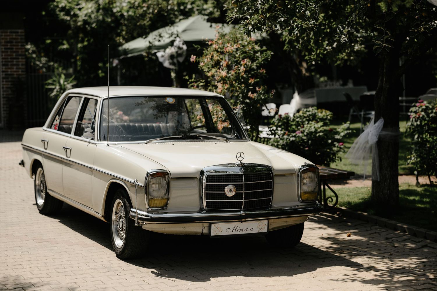 Vintage white Mercedes-Benz sedan parked on a brick driveway surrounded by lush green trees on a sunny day.