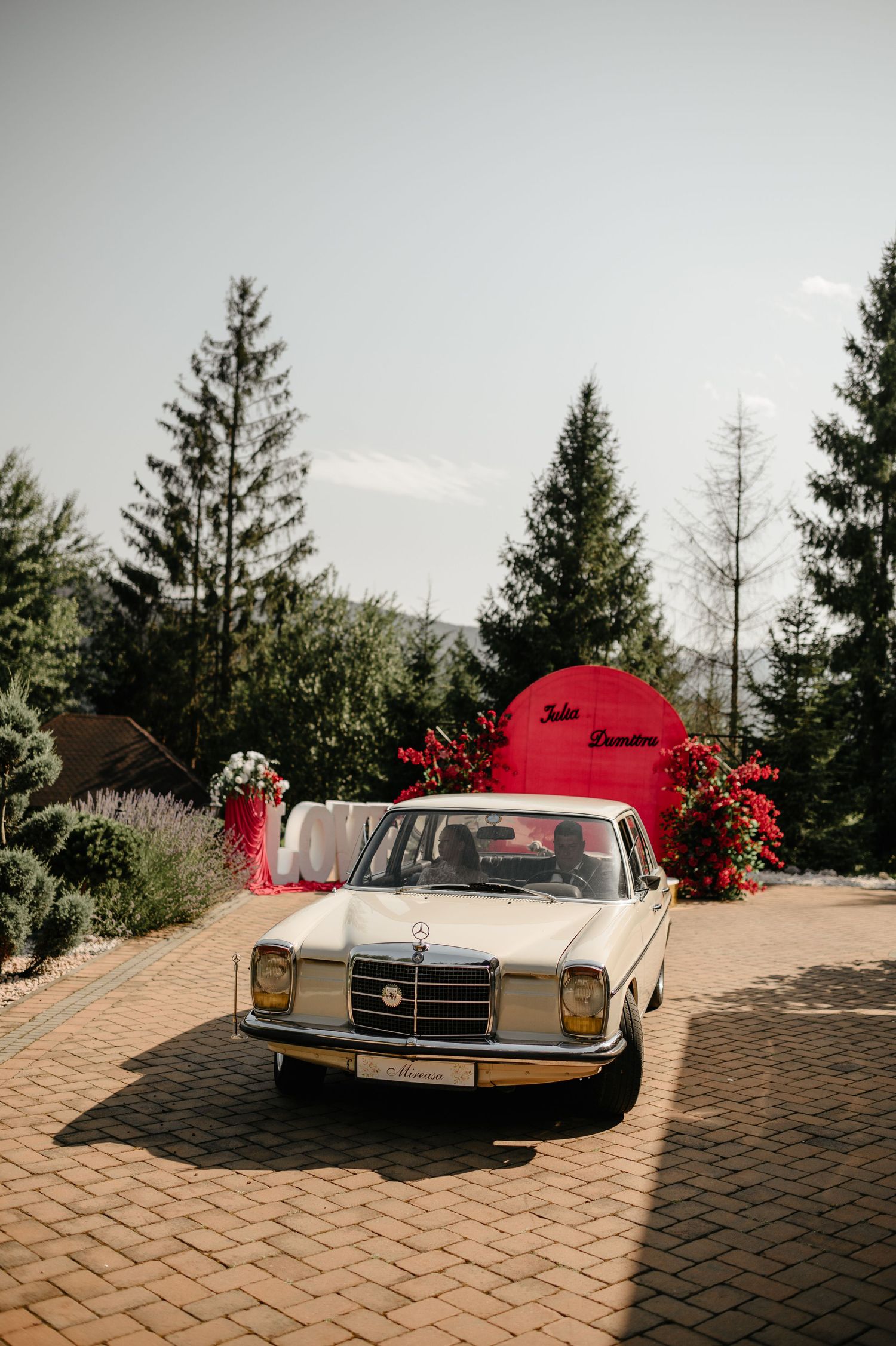 Vintage white Mercedes-Benz parked at wedding venue with red circular backdrop and pine trees.