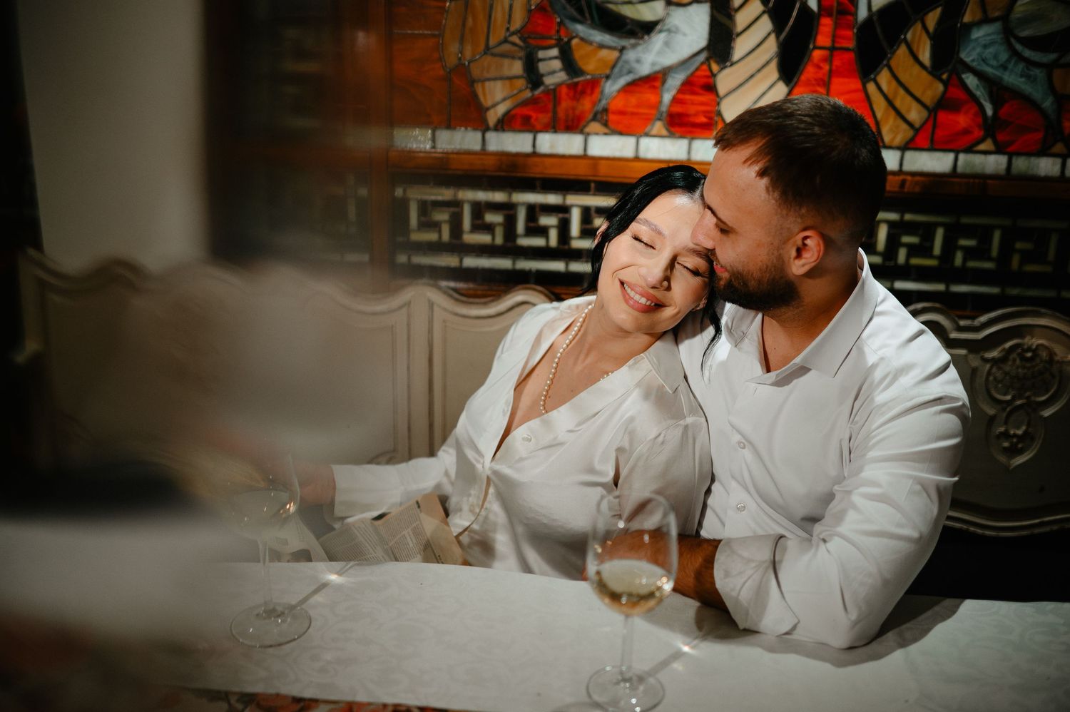 Romantic couple sitting close together at a table with wine glasses near a stained glass window indoors.