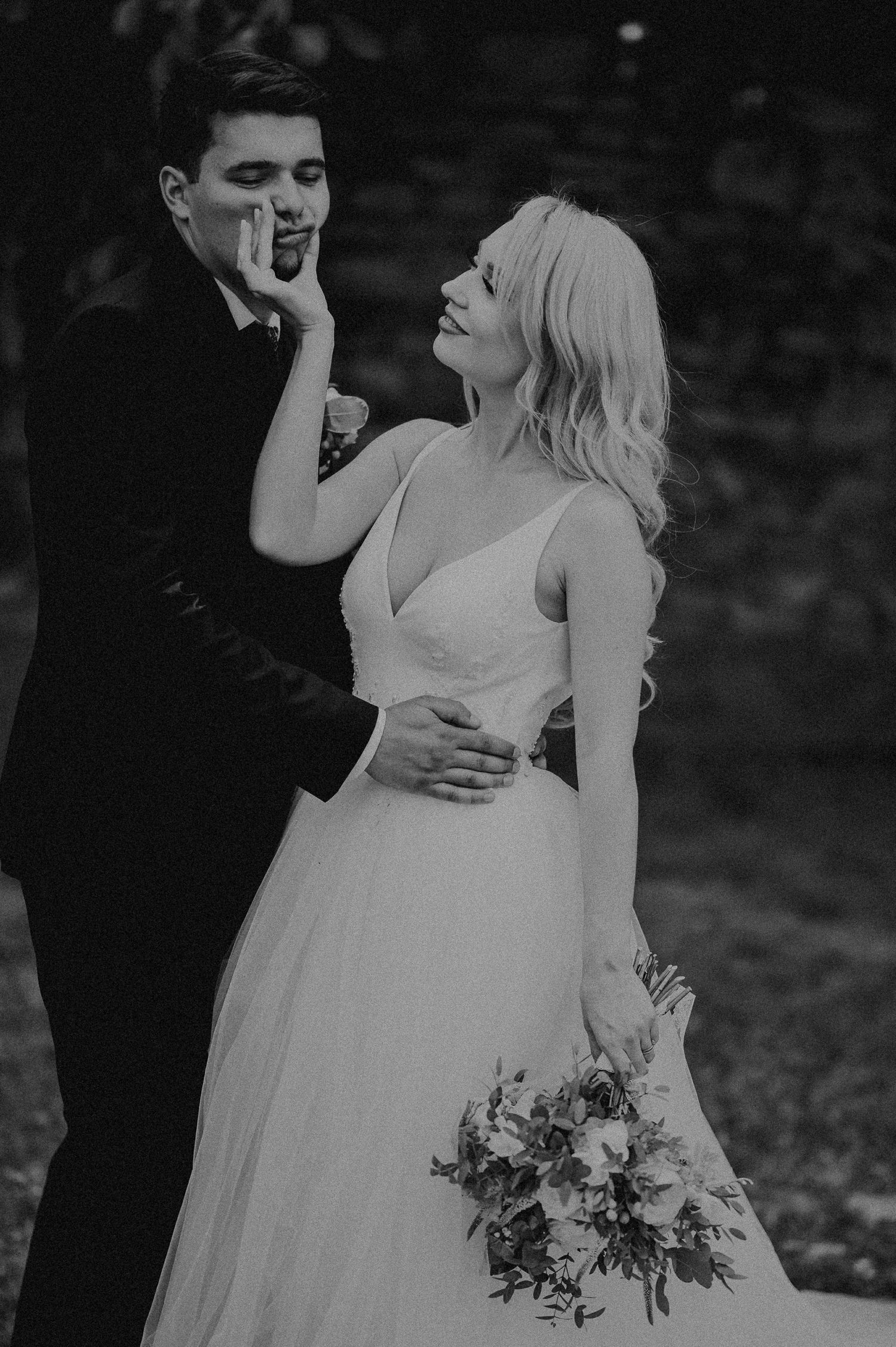 Black and white wedding photo of bride holding bouquet touching grooms face outdoors.