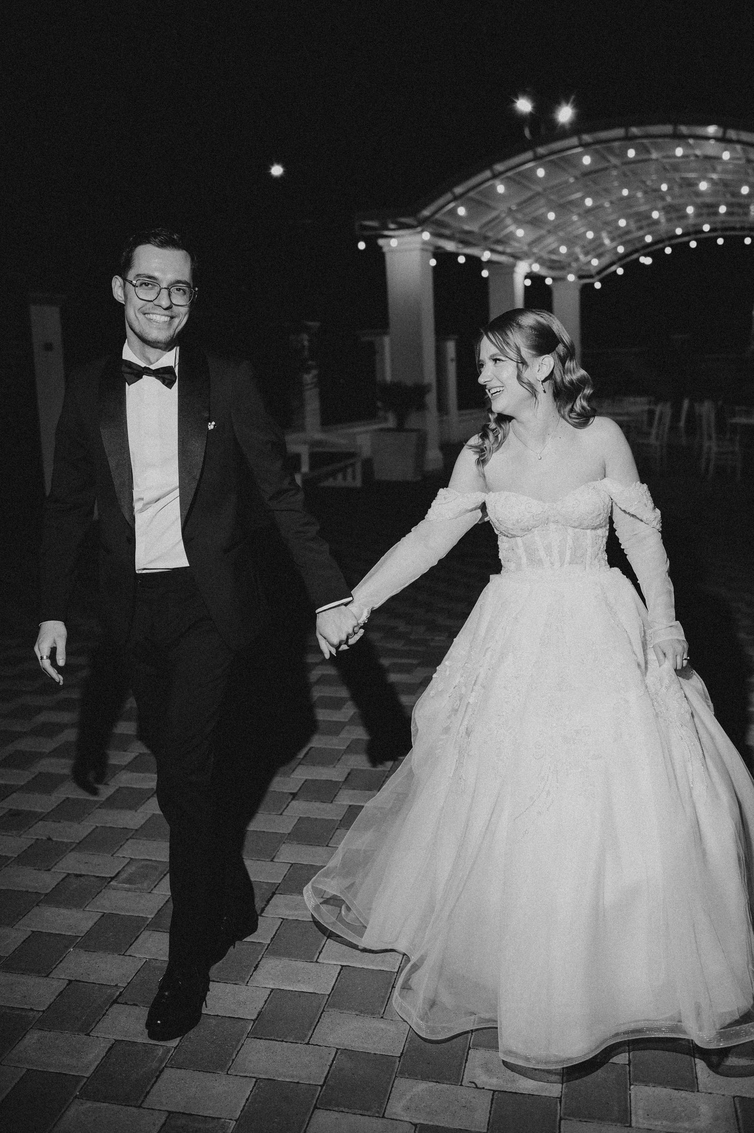 Black and white photo of a bride and groom holding hands, walking at their elegant evening wedding reception.