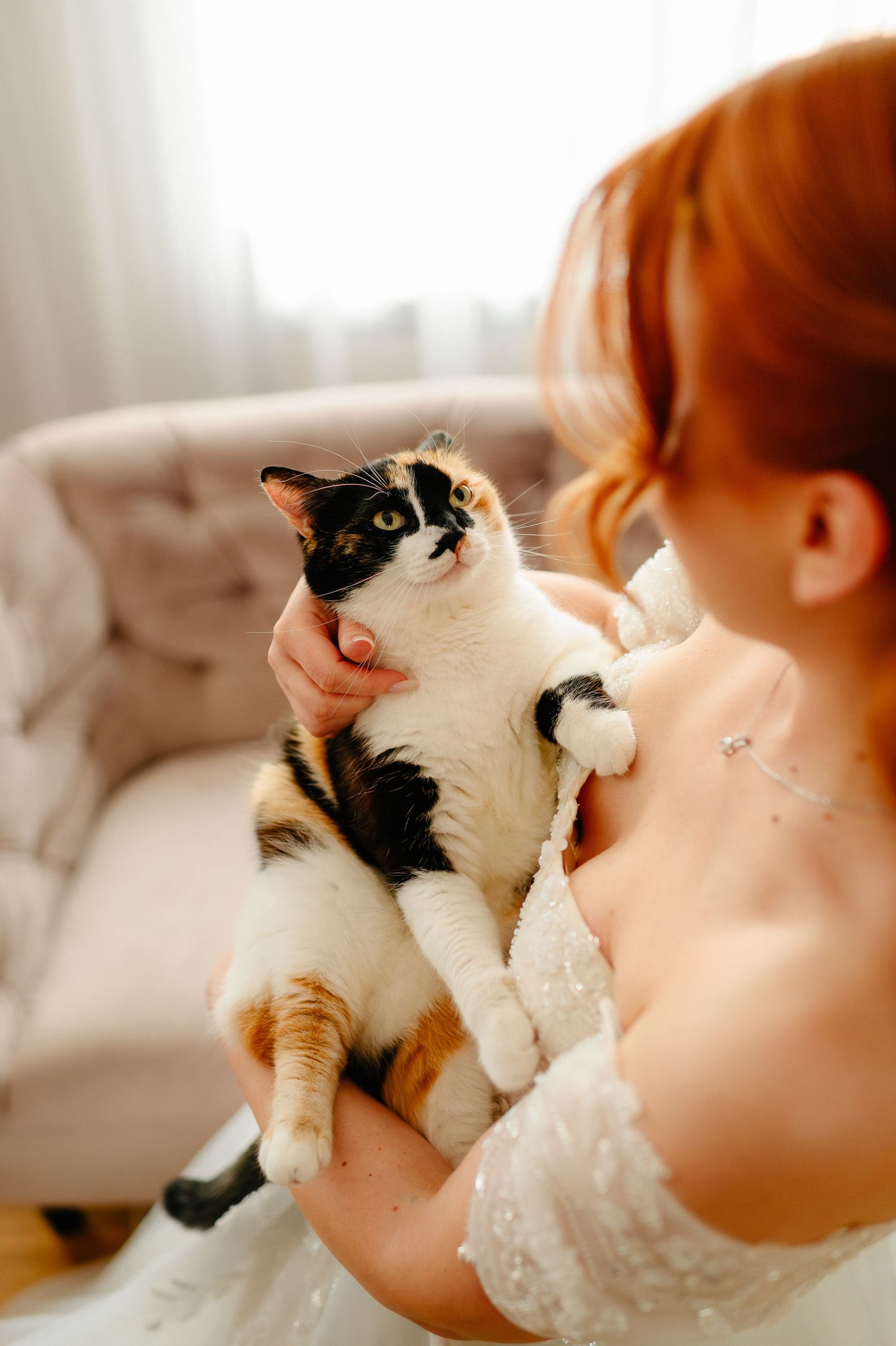 A red-haired bride in a white gown holds a calico cat near a beige sofa in a bright, elegant room.