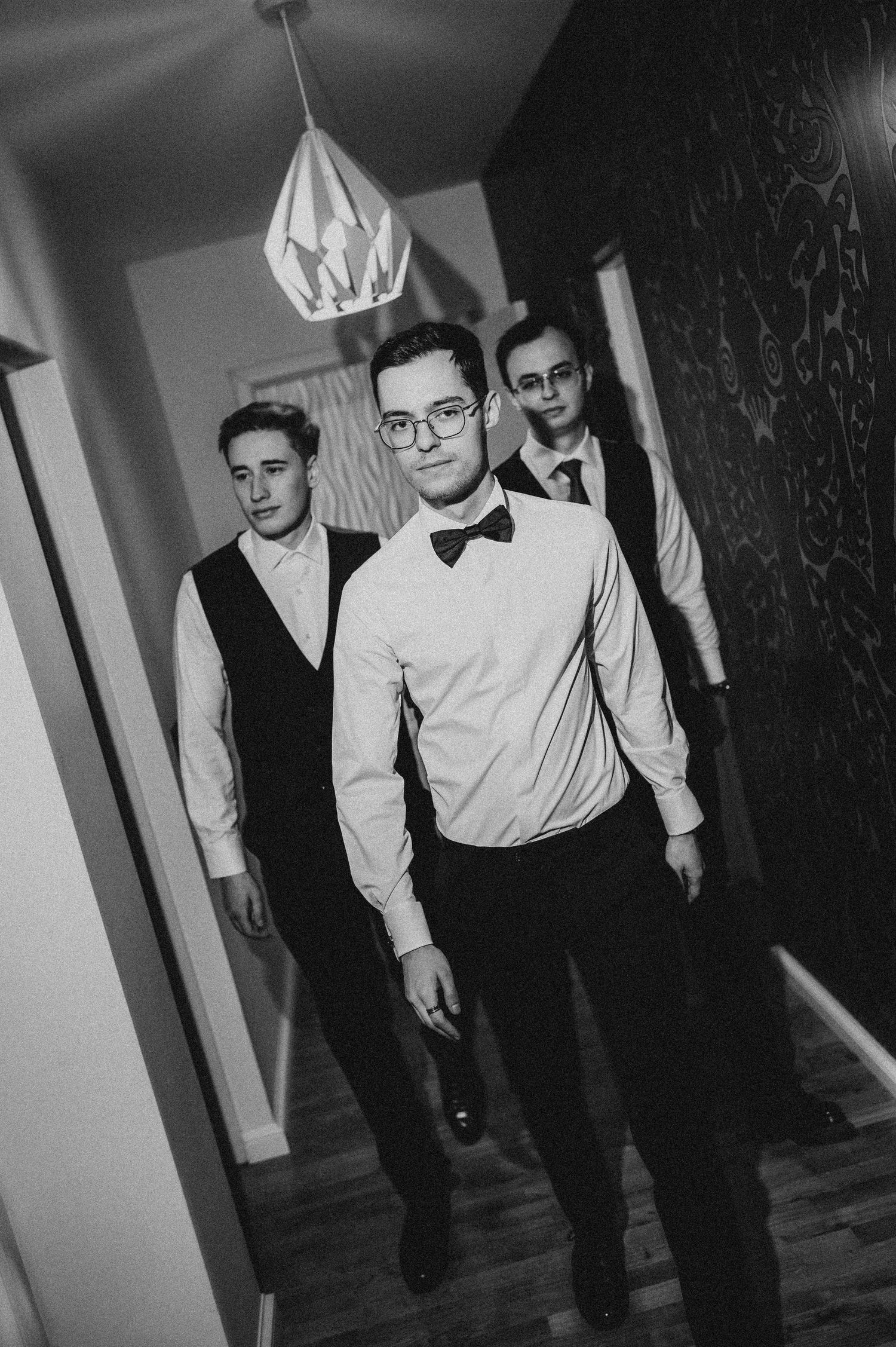 Black and white photo of three young men in formal vests and bow ties walking down a hallway.