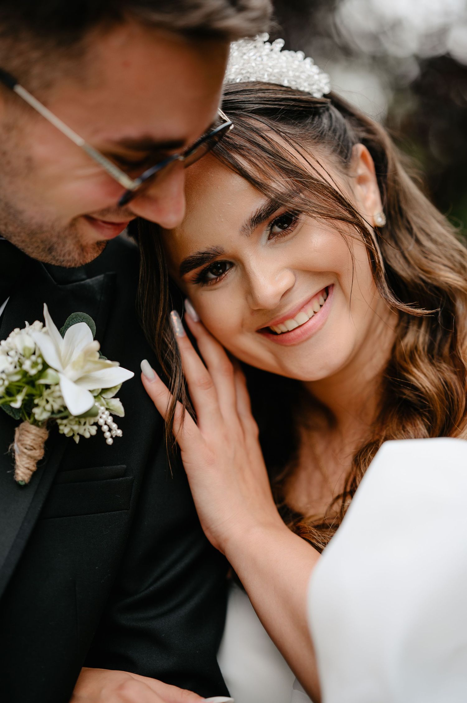 Close-up of smiling bride wearing tiara leaning against groom in black suit with white boutonniere.