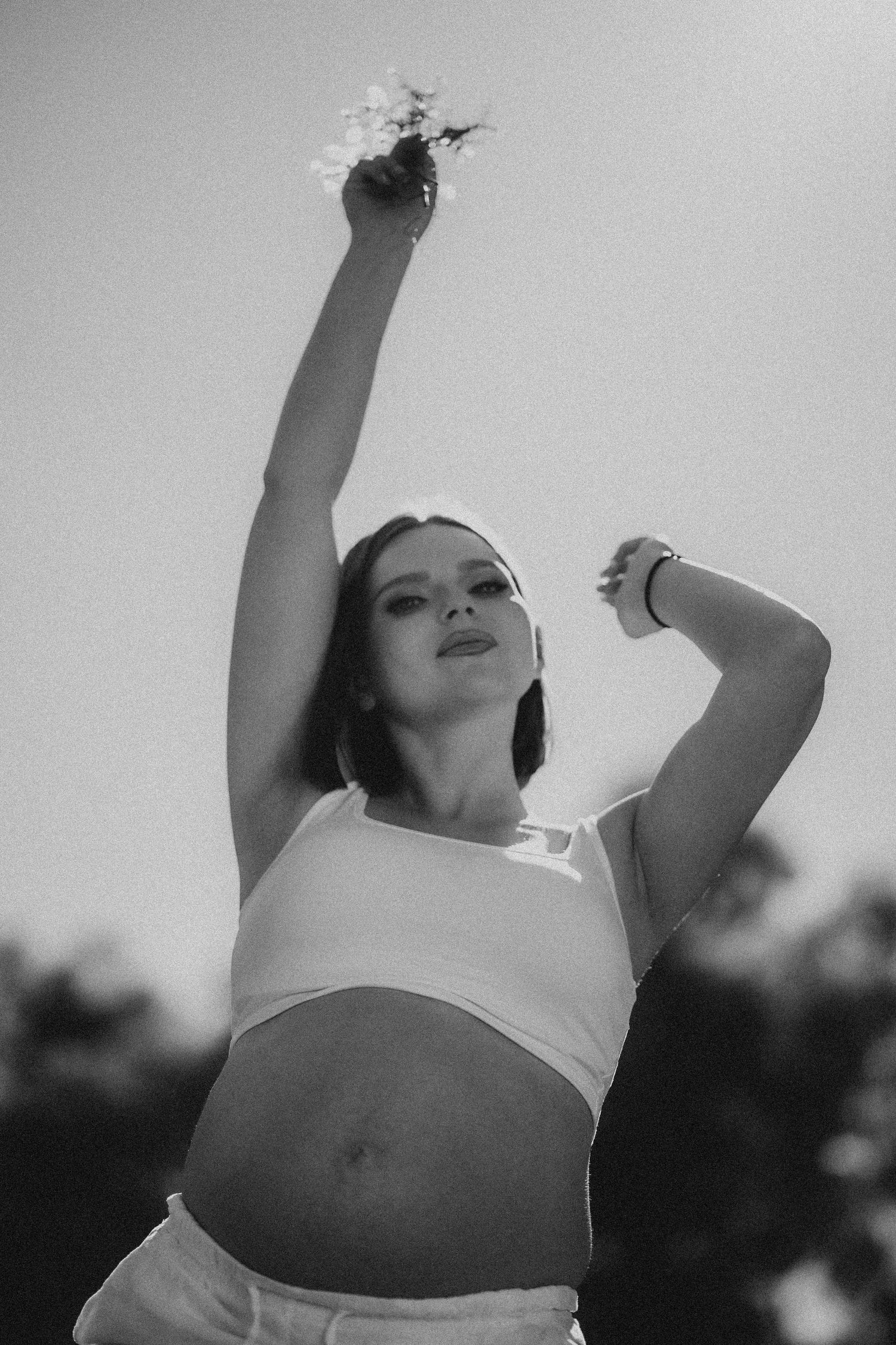 Pregnant woman in white crop top raising arm with flowers in artistic black and white outdoor photo.