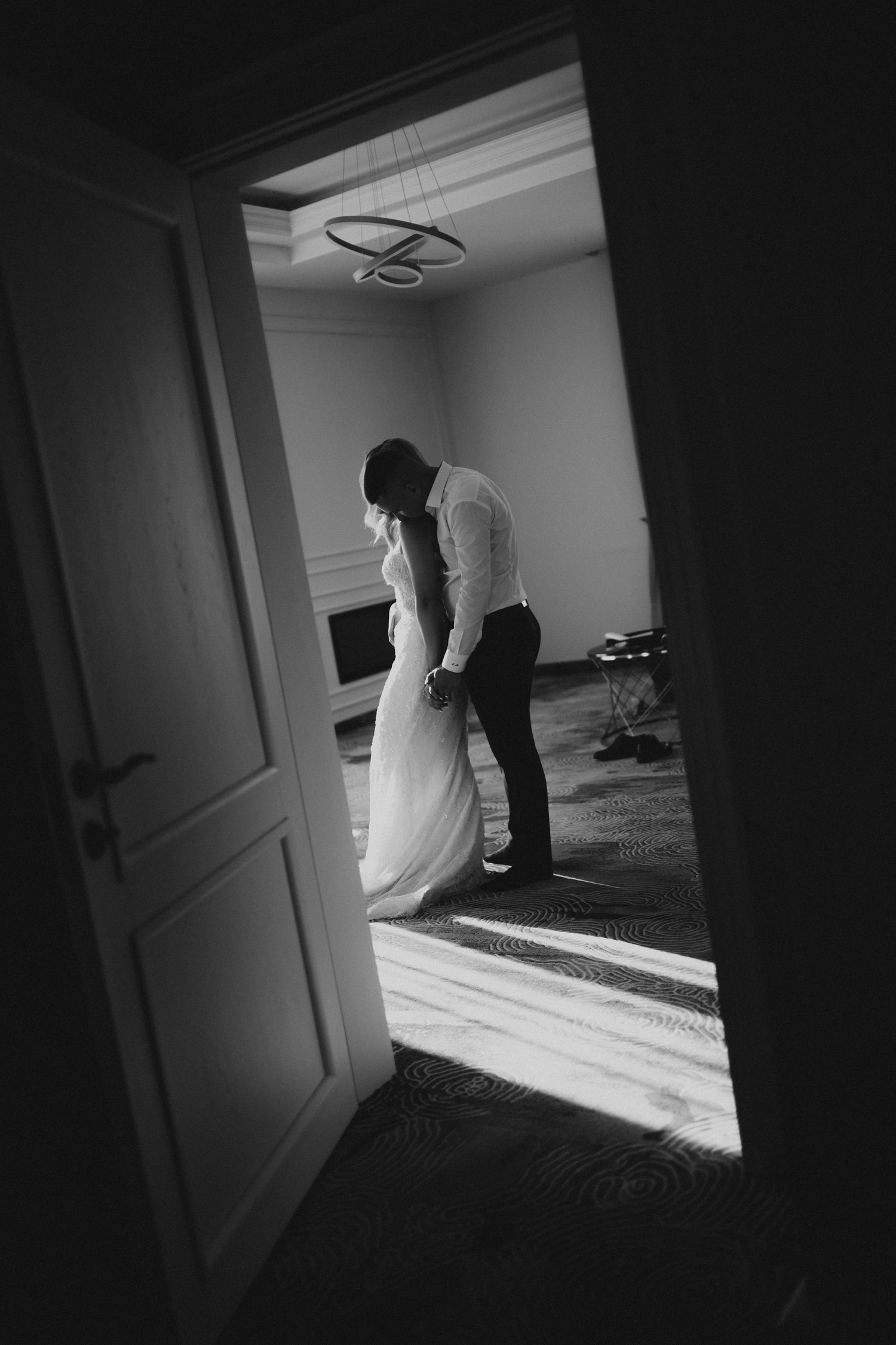 Black and white photo of bride and groom sharing intimate moment in sunlit room, viewed through open doorway.
