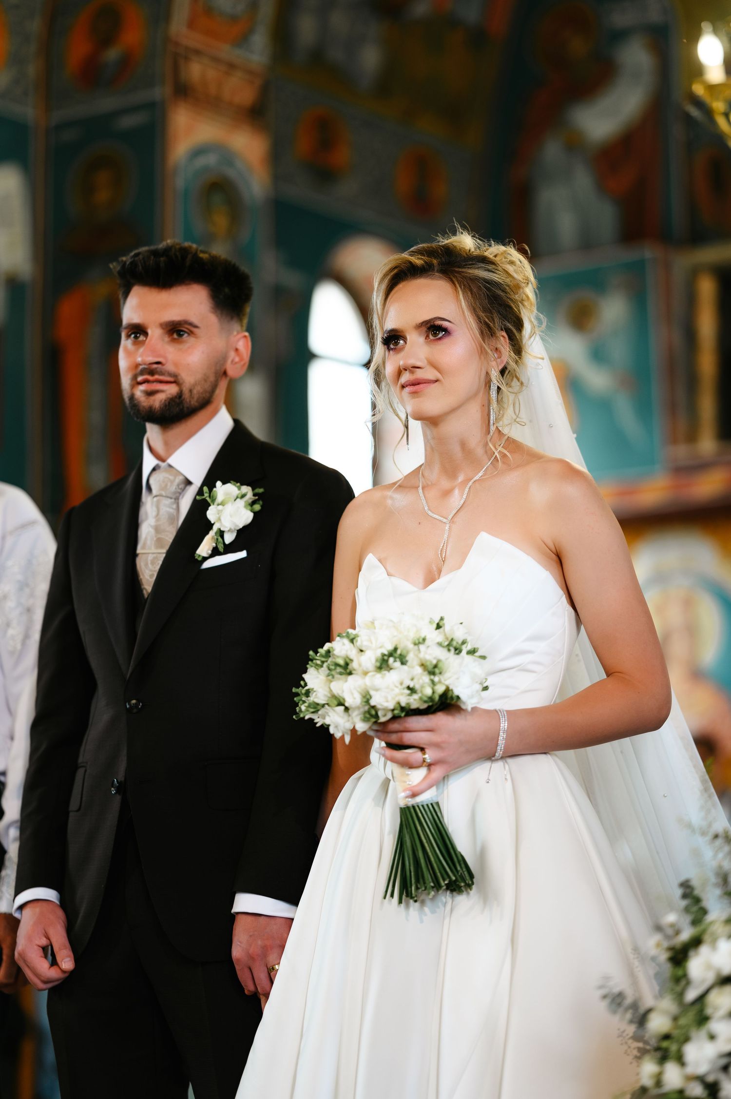 Bride in white strapless gown holding bouquet stands with groom in black suit during Orthodox church wedding ceremony.