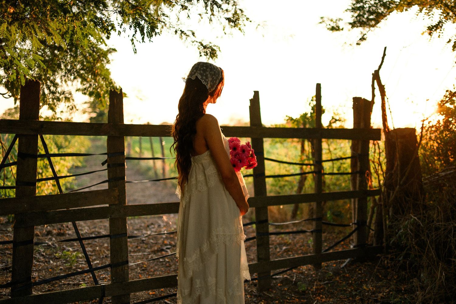 Woman in white dress holding pink flowers stands by rustic wooden fence at golden sunset.