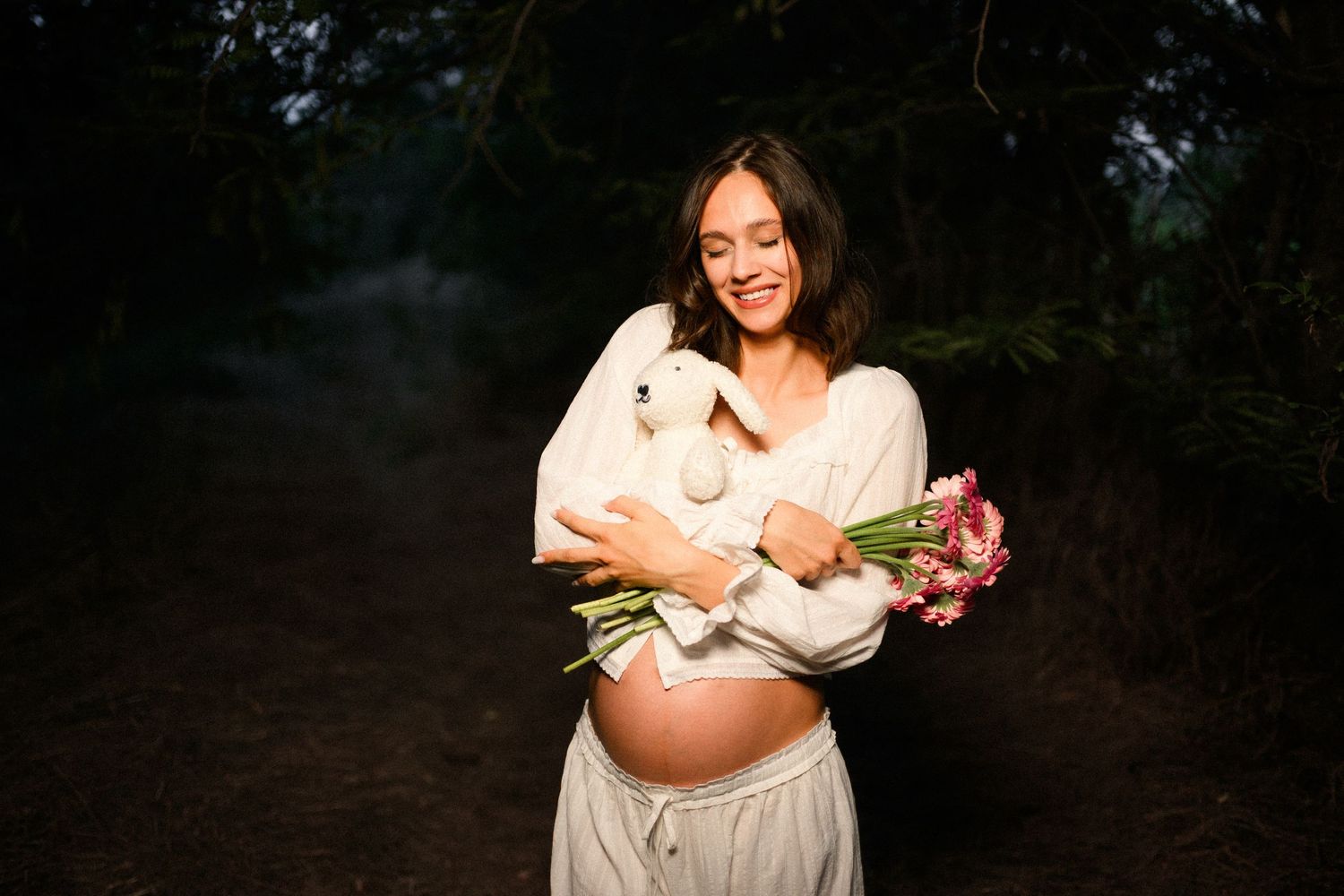 Smiling pregnant woman in white outfit holds flower bouquet outdoors at night, cradling her baby bump in a dark forest setting.