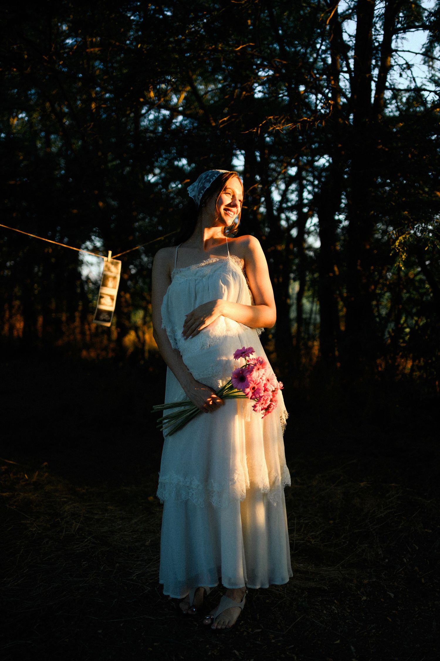 Smiling pregnant woman in white dress holding pink flowers, bathed in golden sunset light in a forest setting.