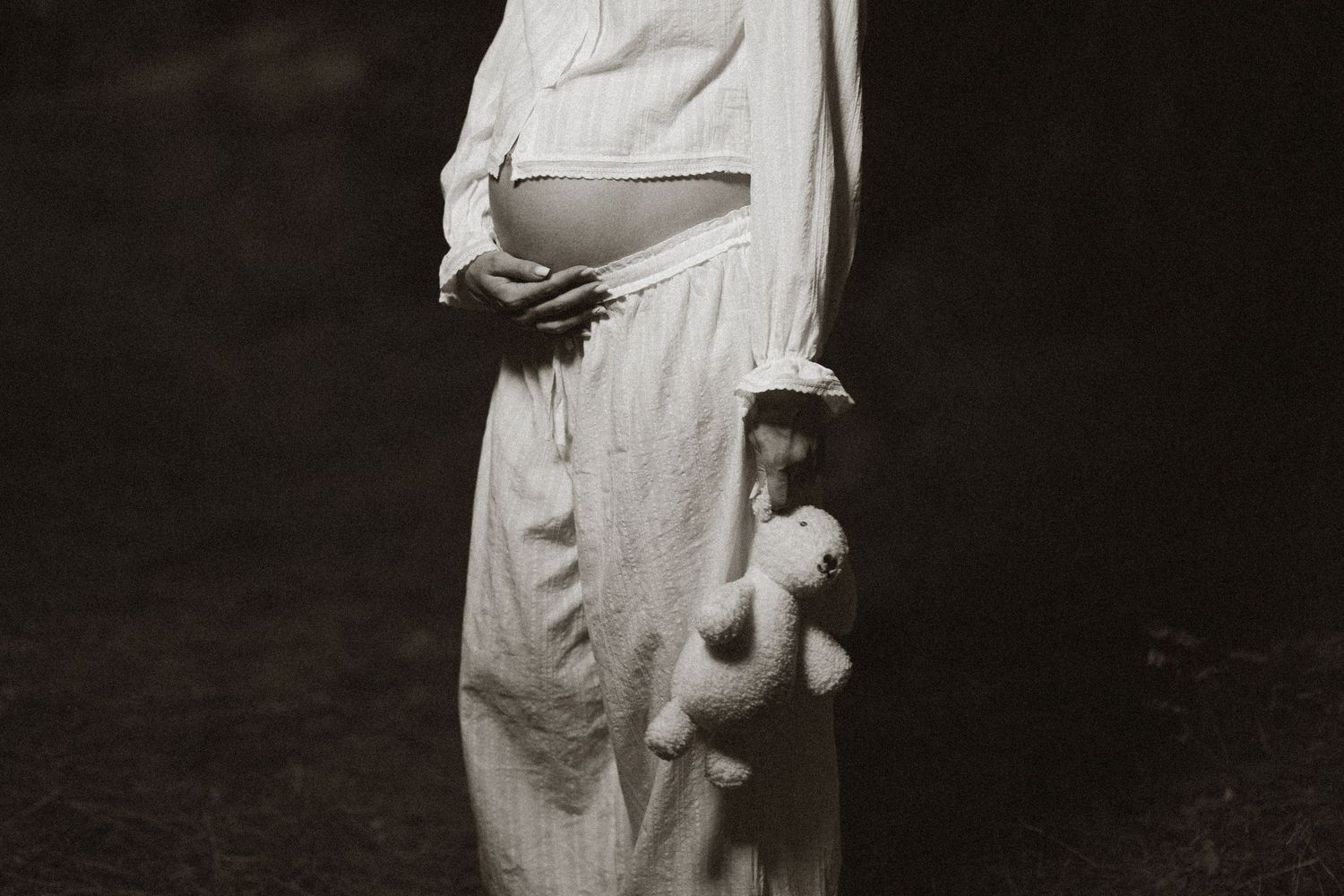 Pregnant woman in white dress holding a teddy bear, black and white maternity photography.