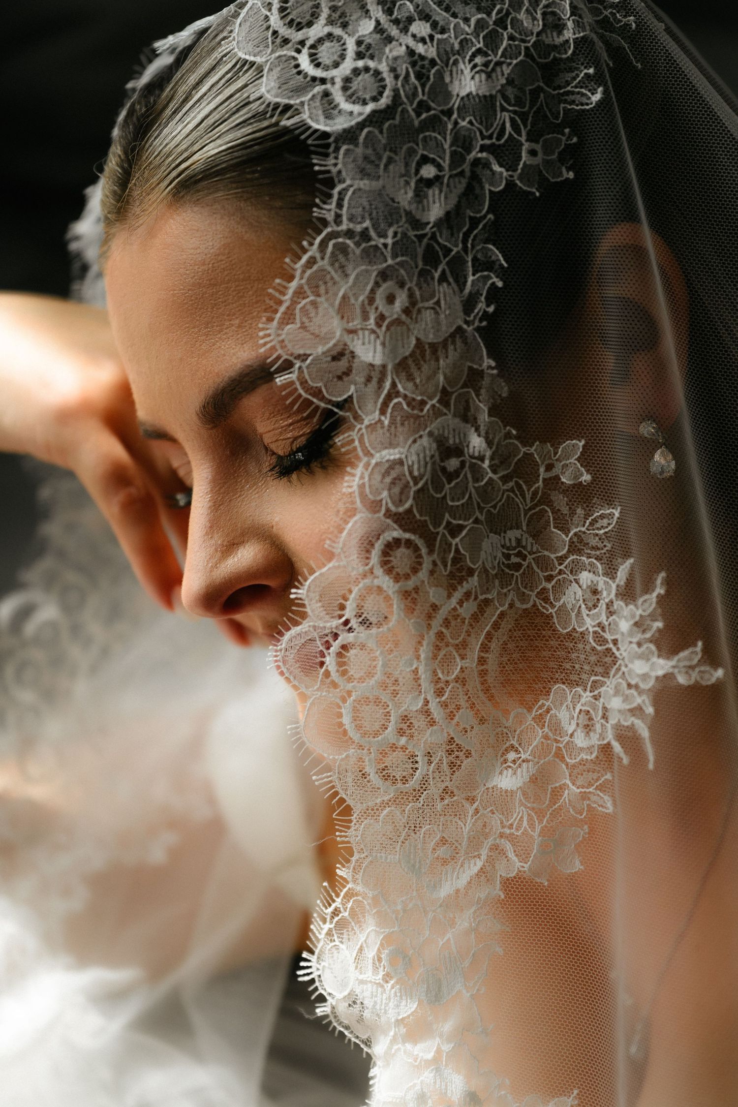 A bride wearing an intricate white lace veil with floral patterns, eyes downcast in an elegant wedding portrait.