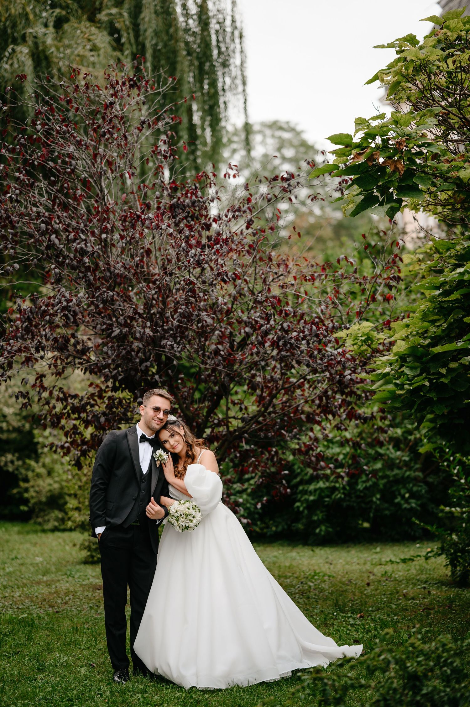 Bride and groom embracing in a lush garden with burgundy trees on their wedding day.