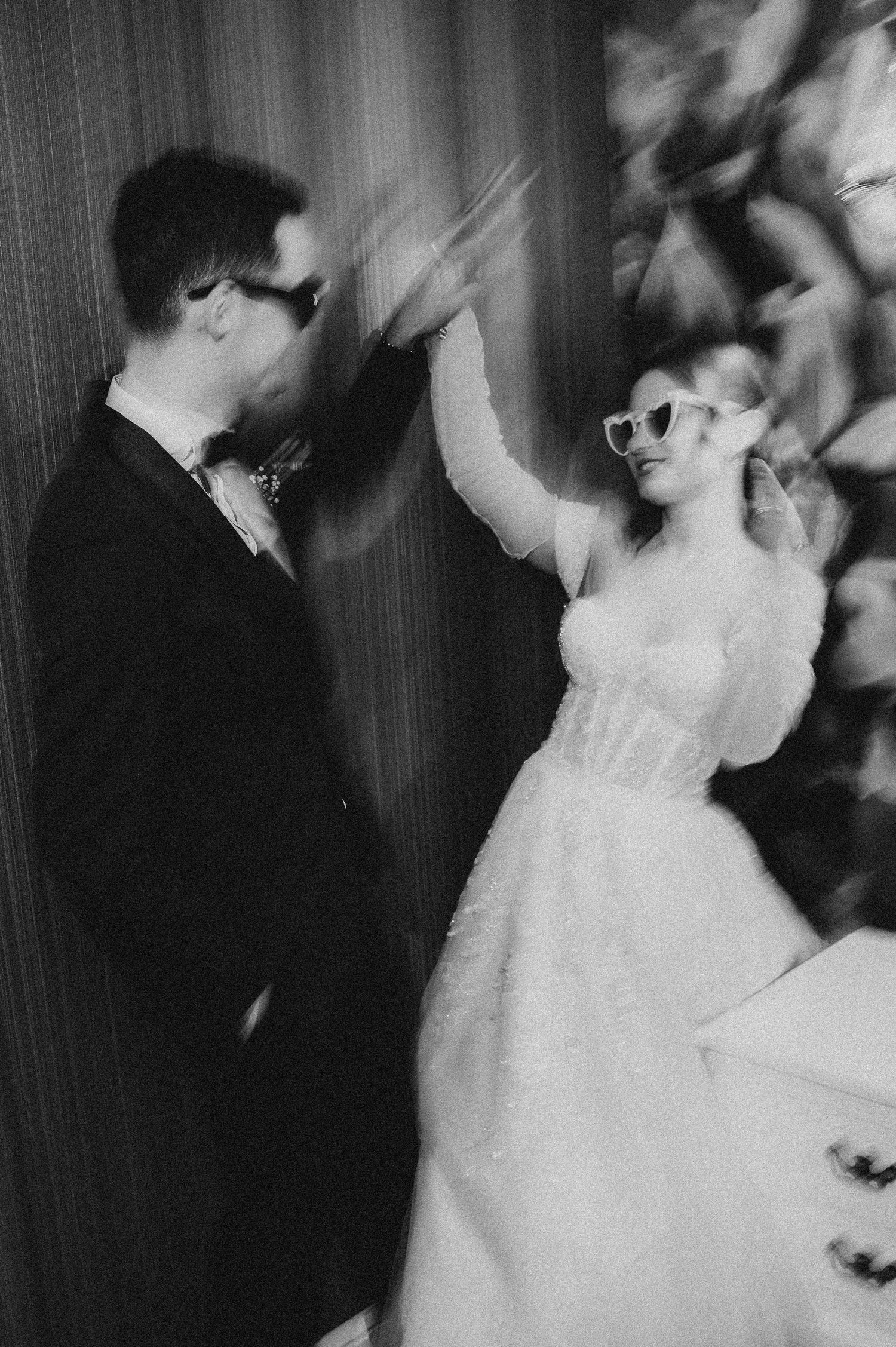 Black and white blurred photo of a bride and groom in sunglasses sharing a joyful dance on their wedding day.