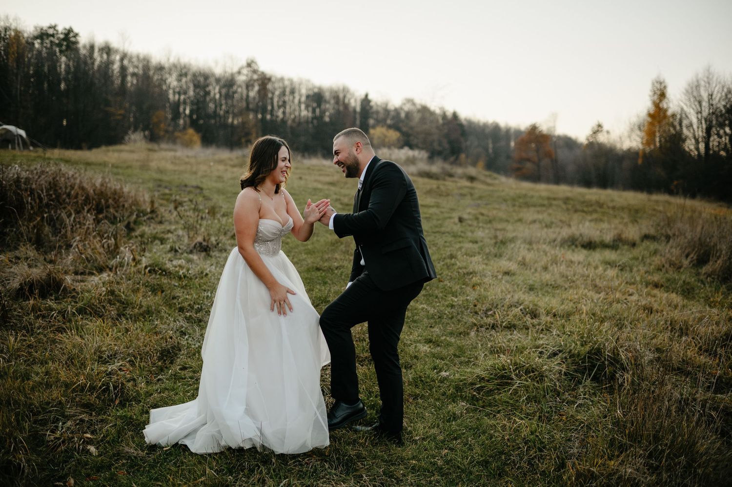 Bride and groom dancing joyfully in a rustic autumn field during golden hour wedding portrait.