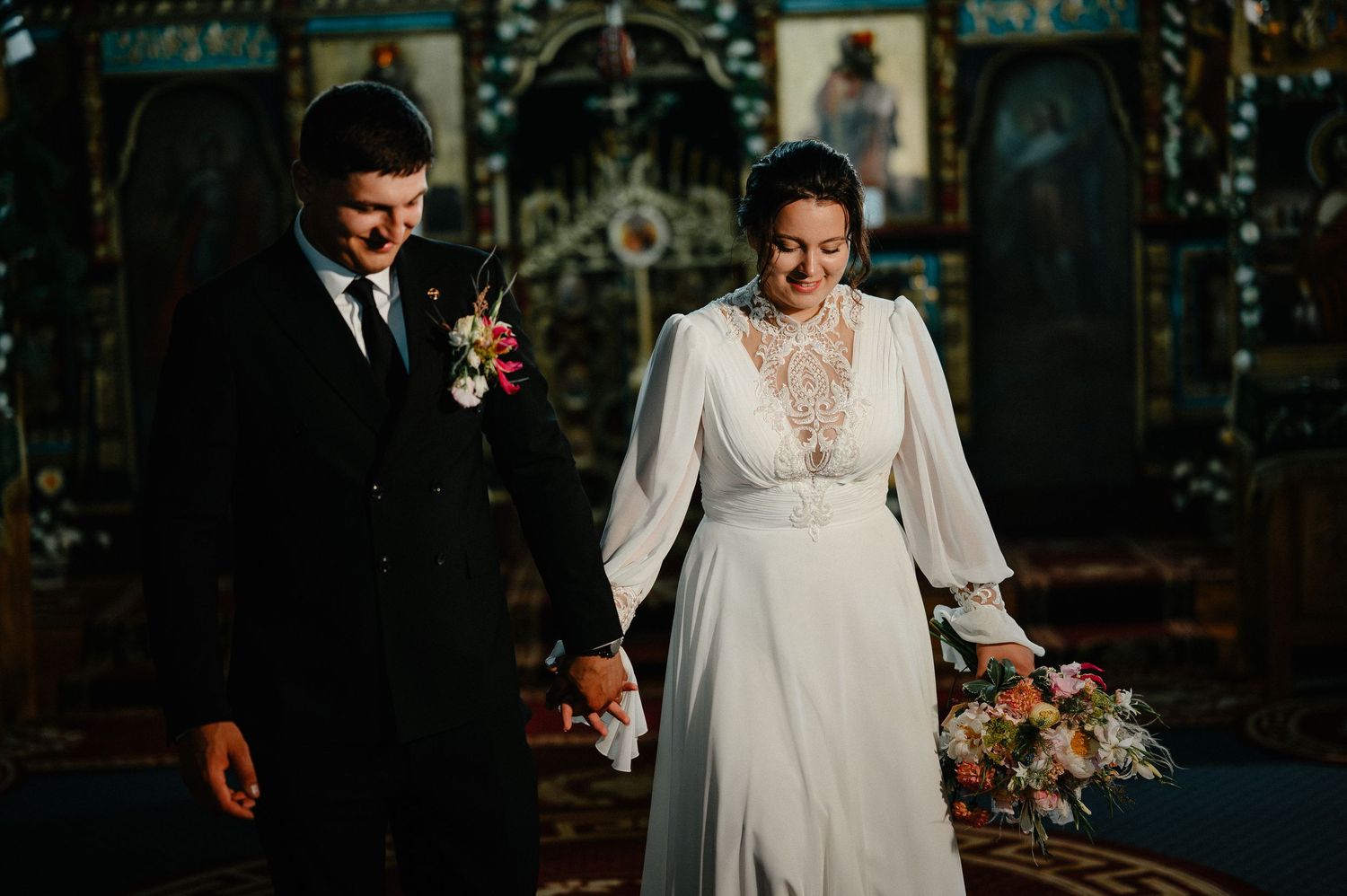 Bride and groom hold hands and smile while walking inside a decorated Orthodox church during wedding ceremony.