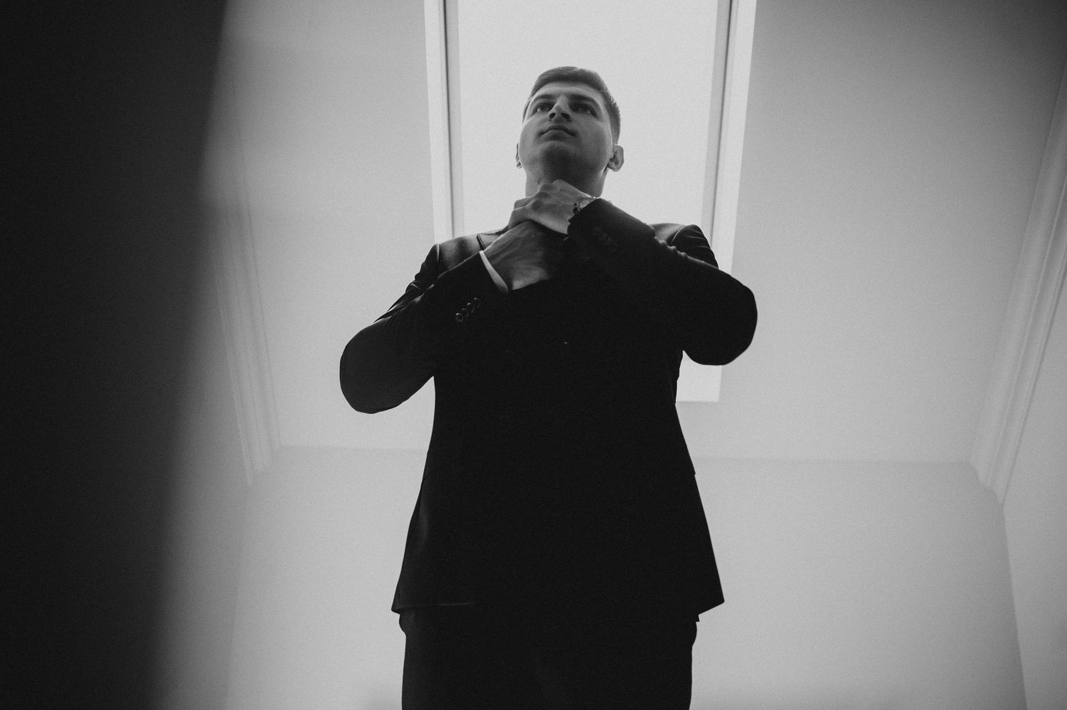 Black and white photo of a groom adjusting his tie while standing in front of a bright window.