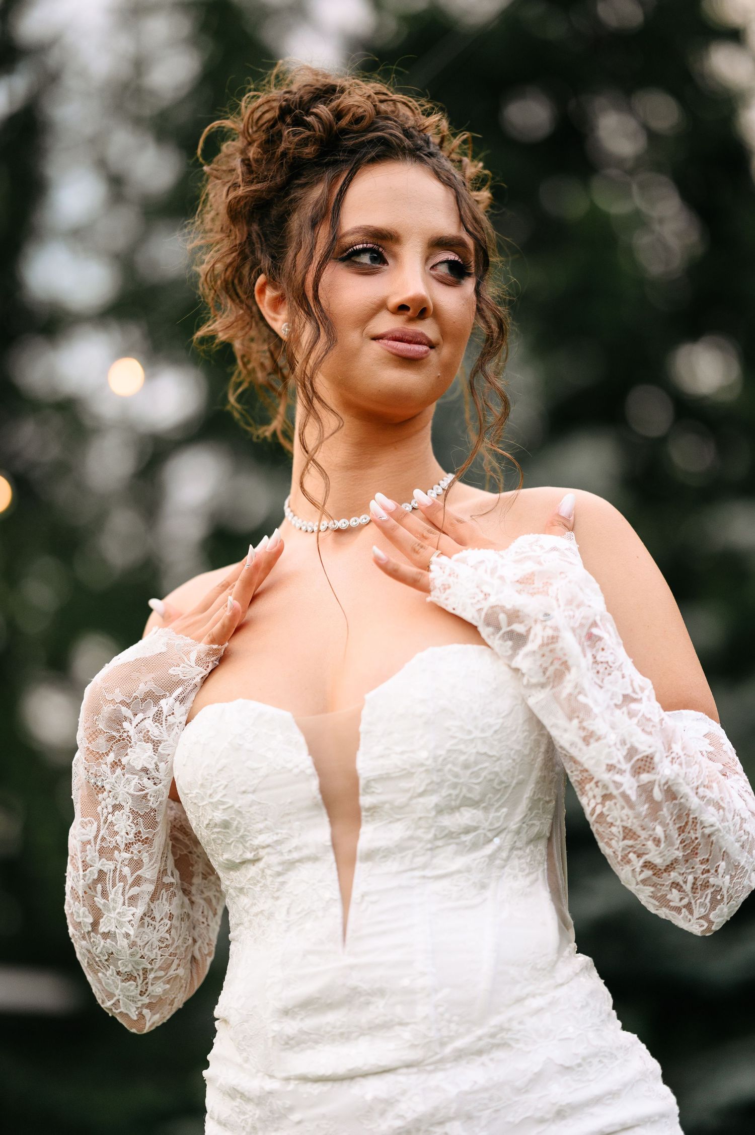 Elegant bride in white lace gown with deep V-neckline, pearl necklace, and updo hairstyle posing outdoors.