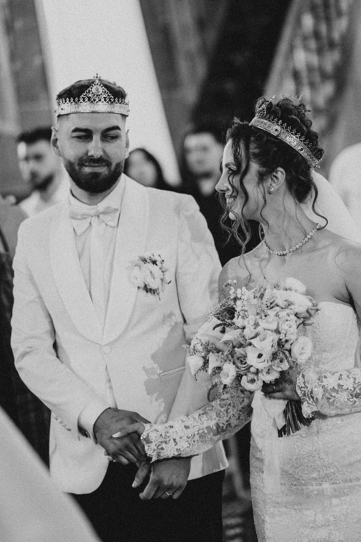 Black and white photo of a bride and groom wearing crowns, holding hands at their wedding ceremony.