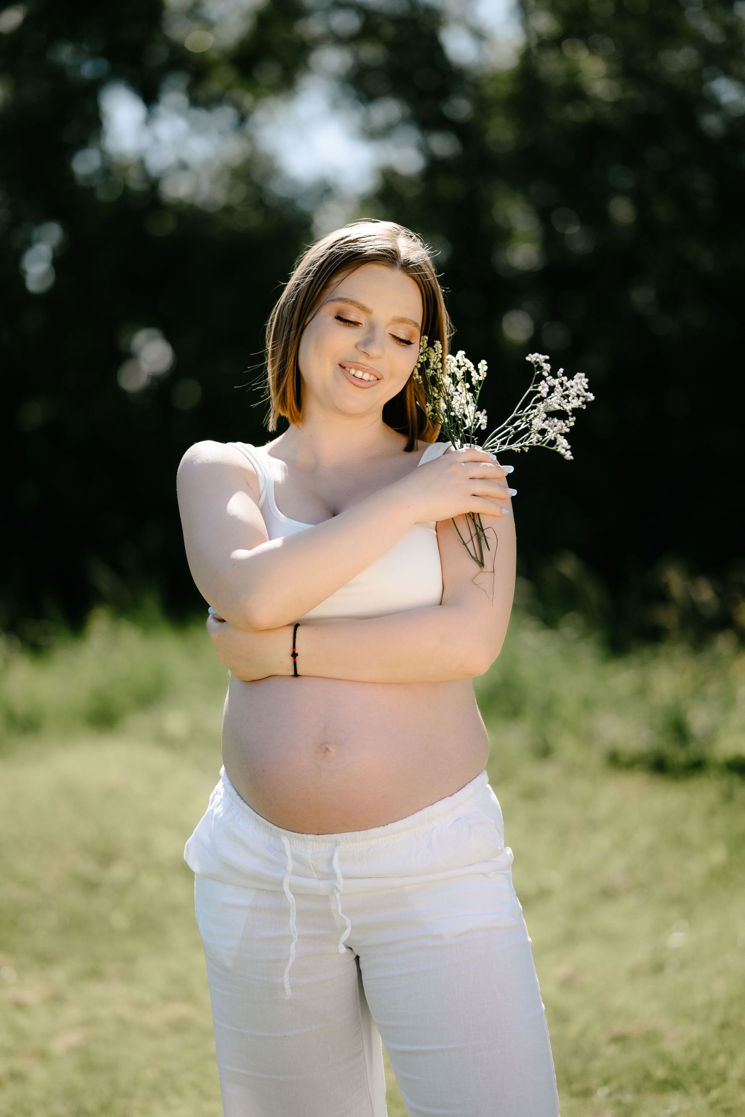 Smiling pregnant woman in white outfit holding wildflowers in sunny outdoor maternity portrait.