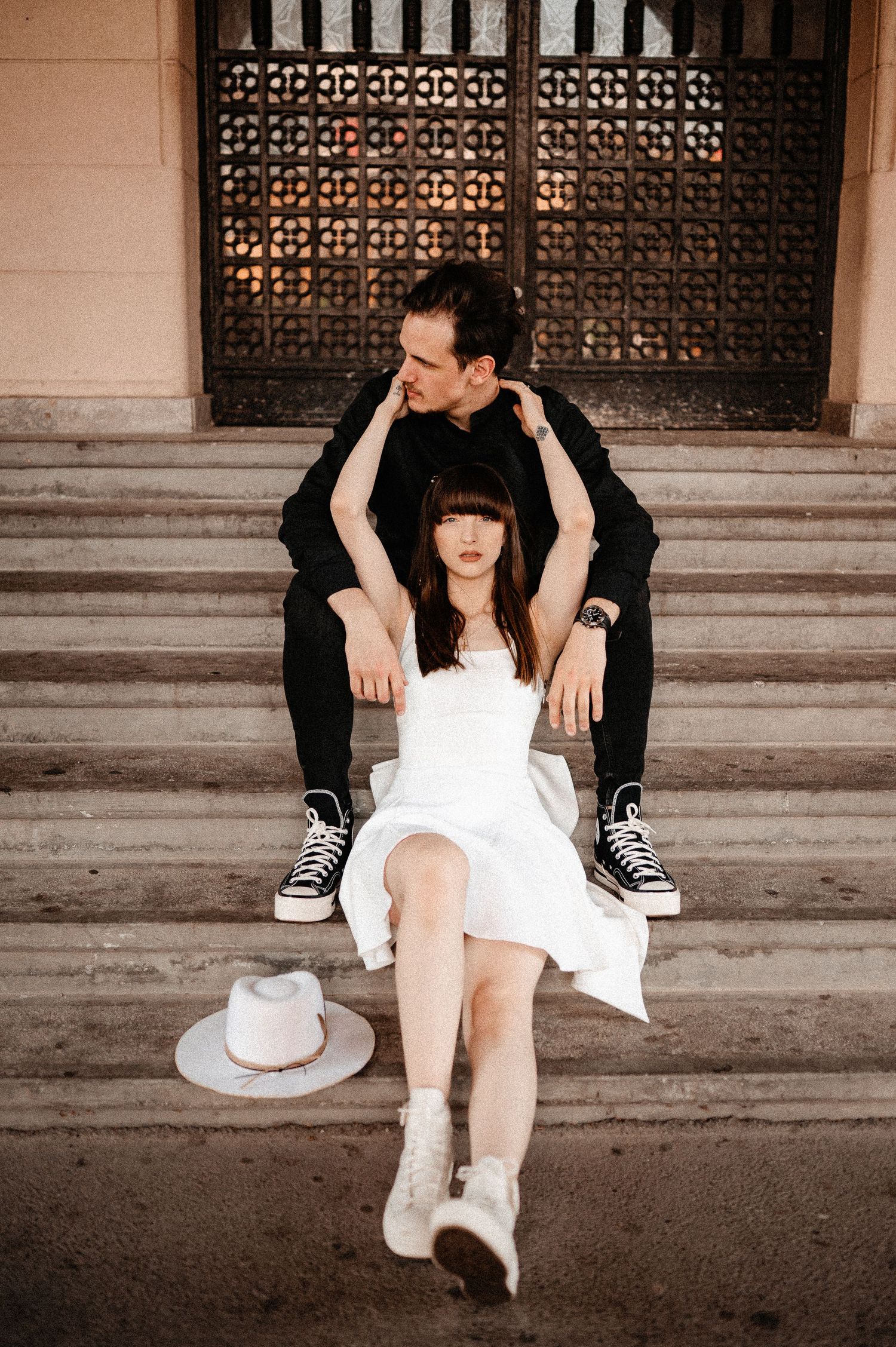 Couple sitting on outdoor steps, man embracing woman in white dress with hat beside them.