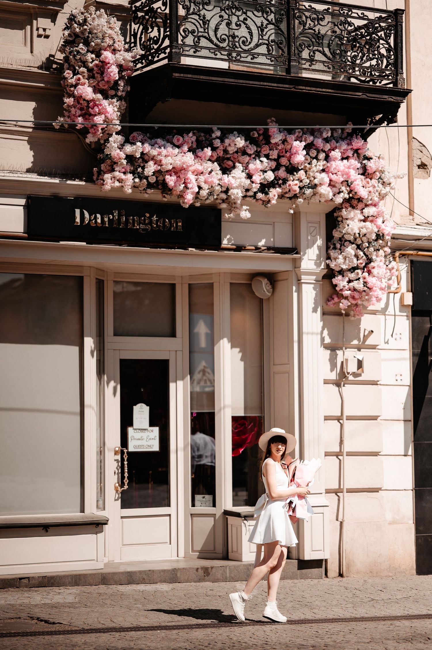 Young woman in white dress and hat standing in front of a flower-adorned Darlington boutique storefront.