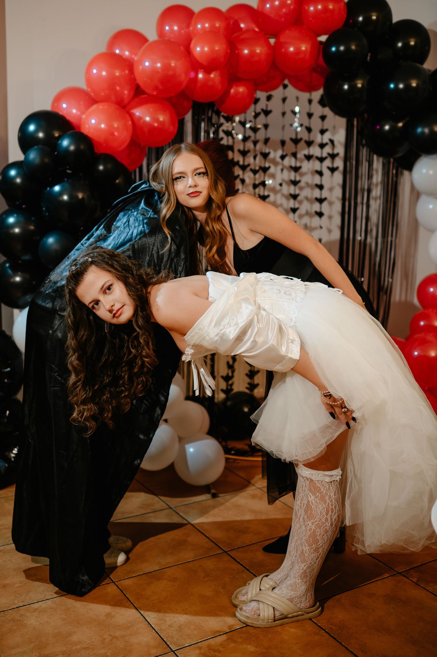 Two women in Halloween costumes pose dramatically before a red and black balloon arch at a party.