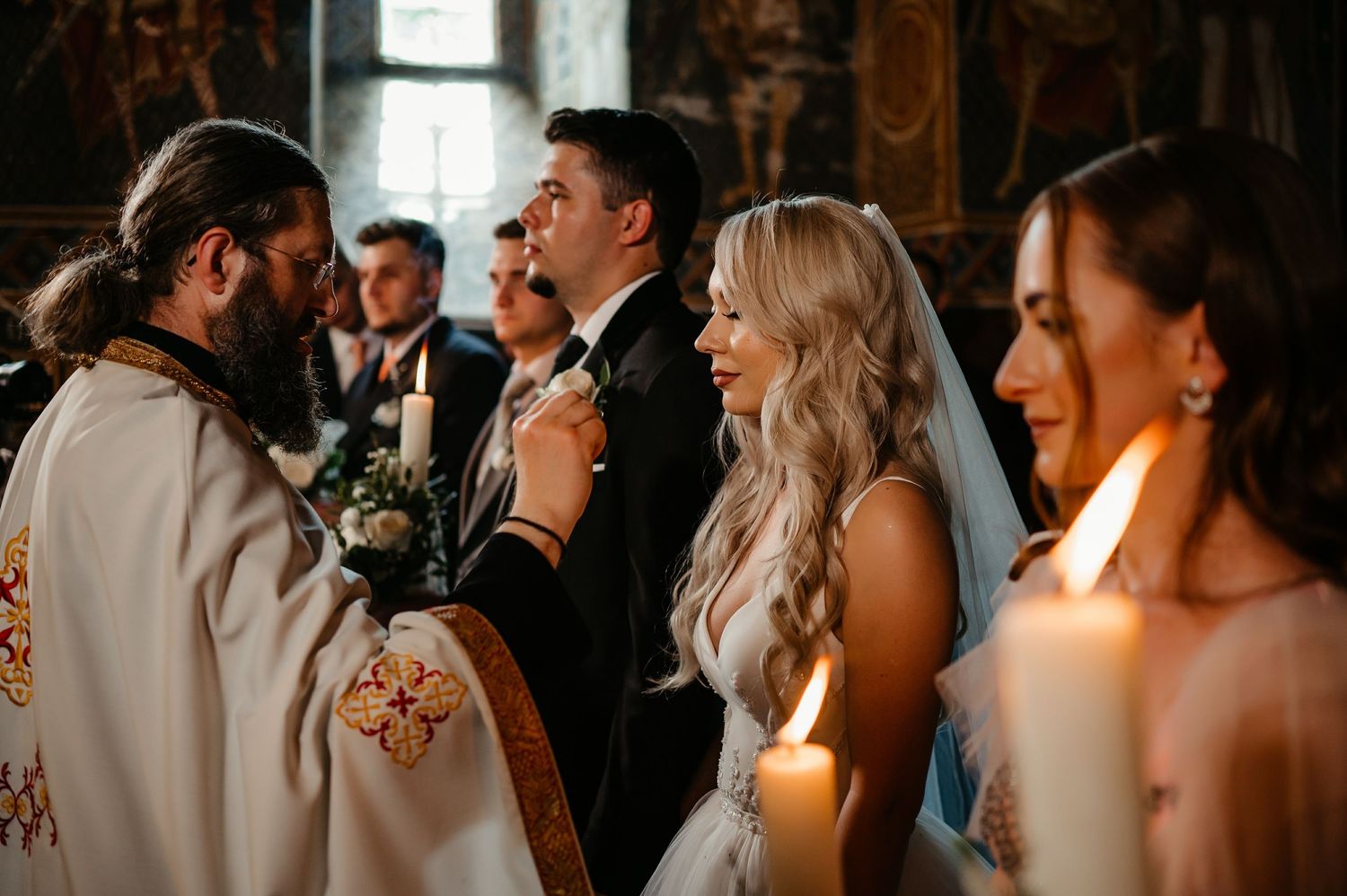 Orthodox priest performing wedding ceremony with candles as bride smiles at groom in decorated church.