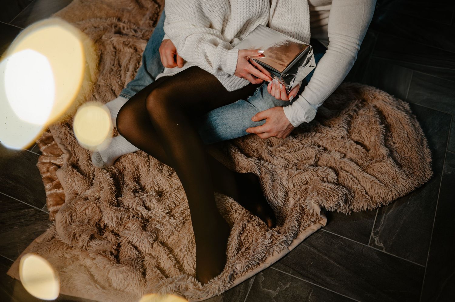 Woman sitting on a fur rug holding a gift box with bokeh lights in the foreground.